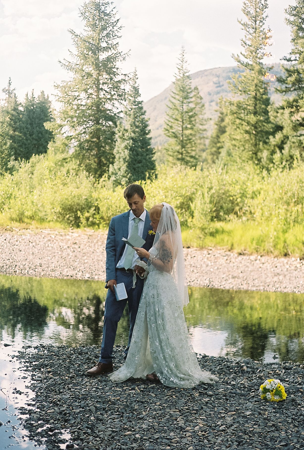 Bride and groom reading vows together on rocky riverbank surrounded by lush mountain scenery.
