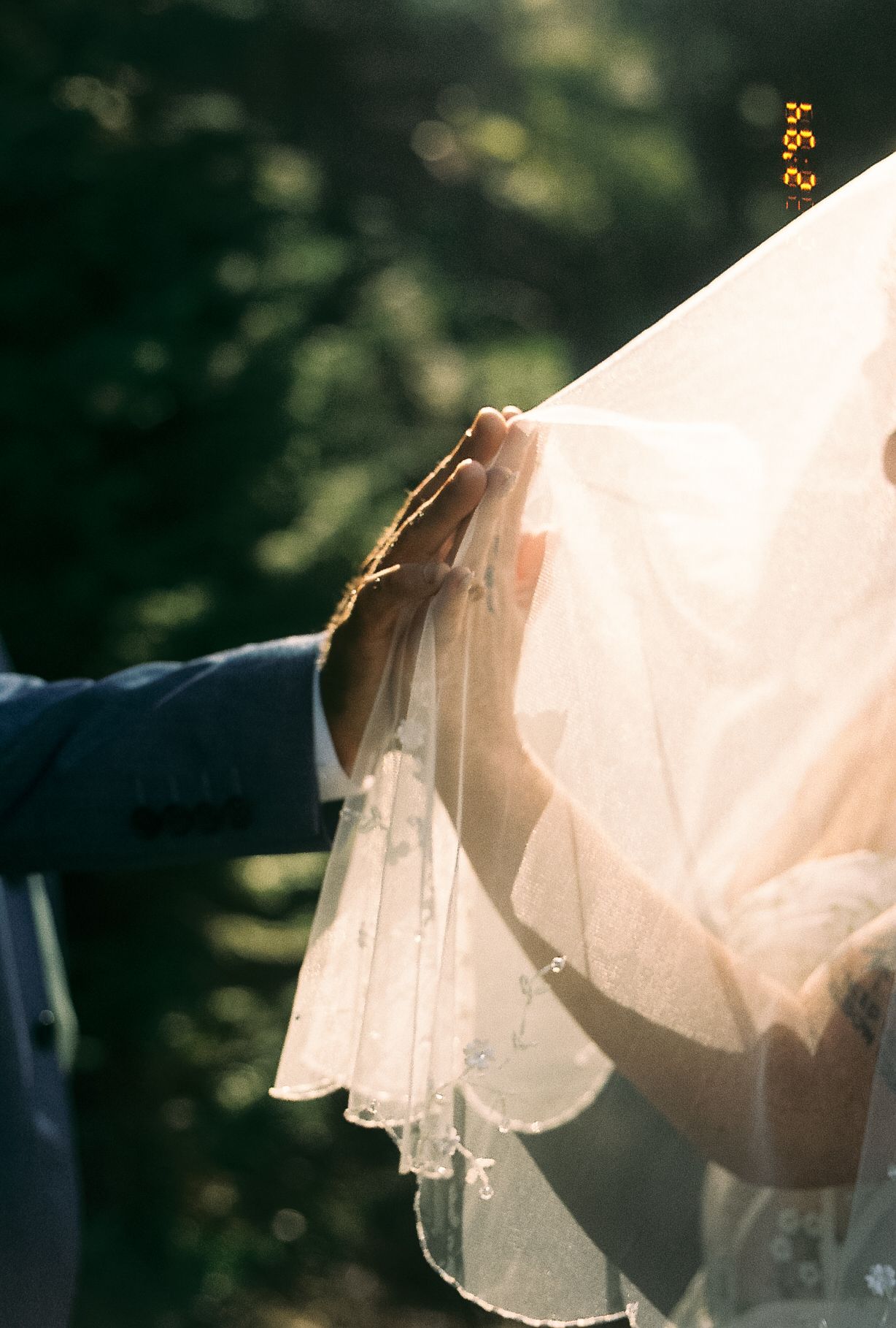 Close-up of groom's hand gently lifting bride's delicate white lace wedding veil in sunlit garden setting.