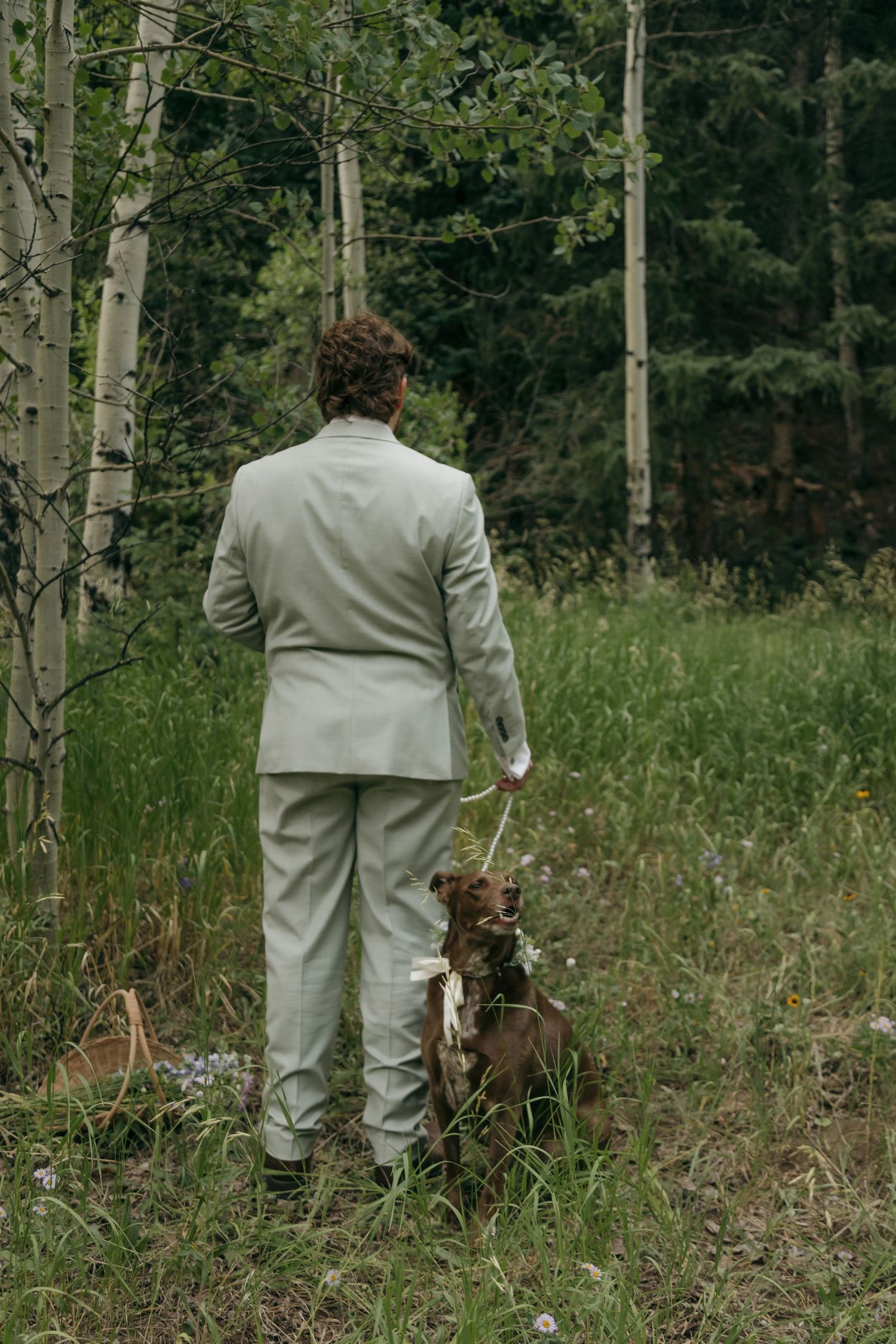 A groom in a light gray suit stands with his brown dog on a leash in a lush forest clearing.