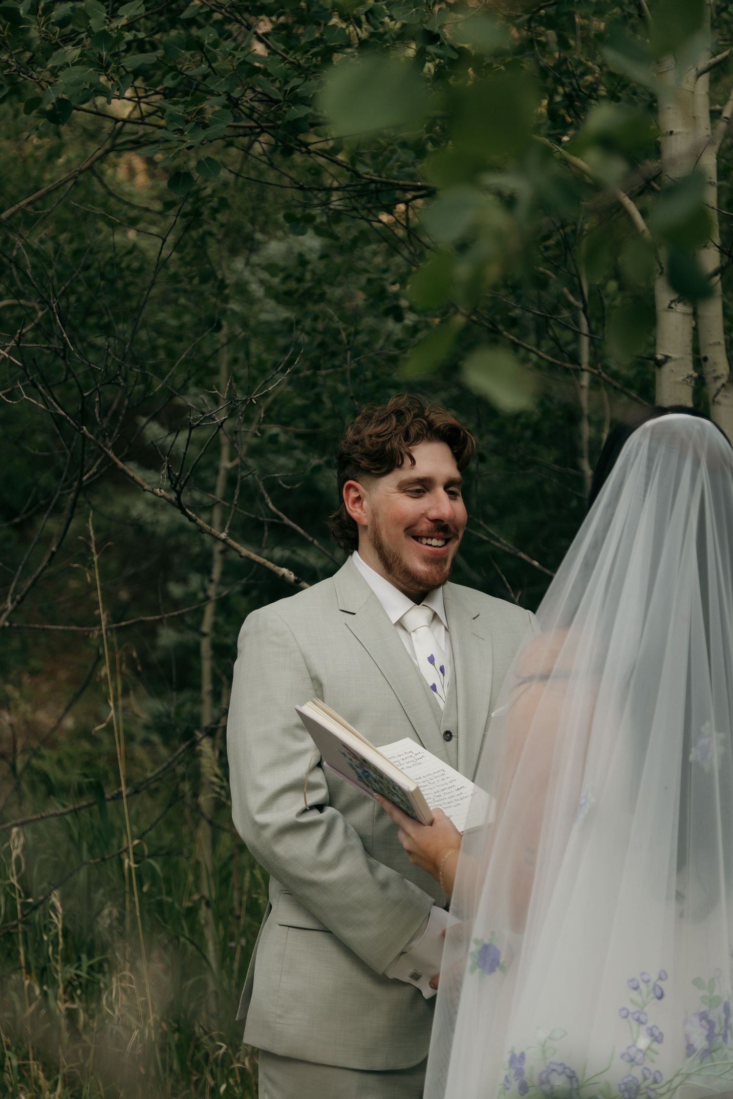 Smiling groom in sage suit holding vows book during outdoor forest wedding ceremony.