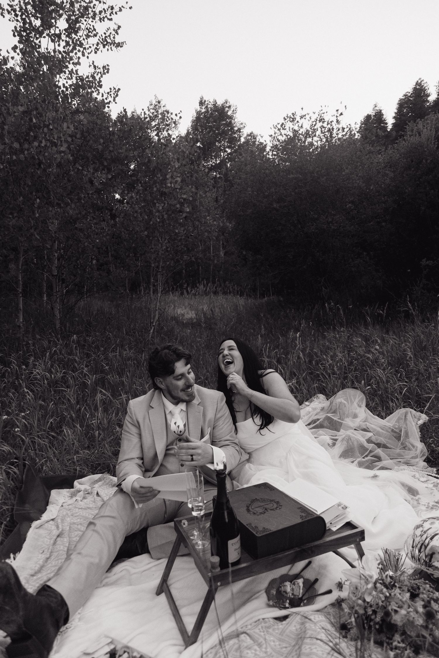 Black and white photo of laughing bride and groom enjoying picnic in meadow.
