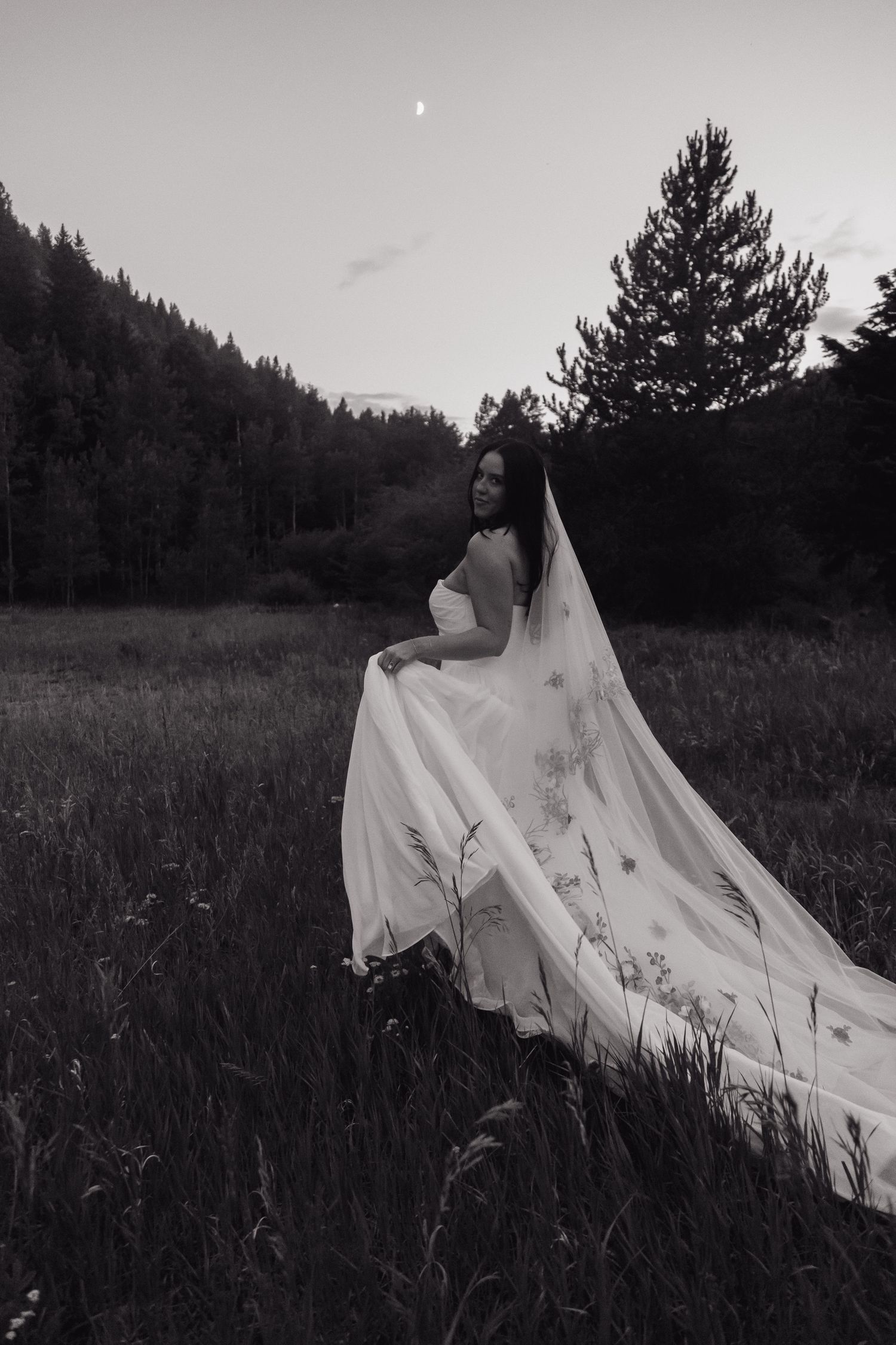 Black and white photo of bride in floral wedding gown with veil flowing in forest meadow at dusk.