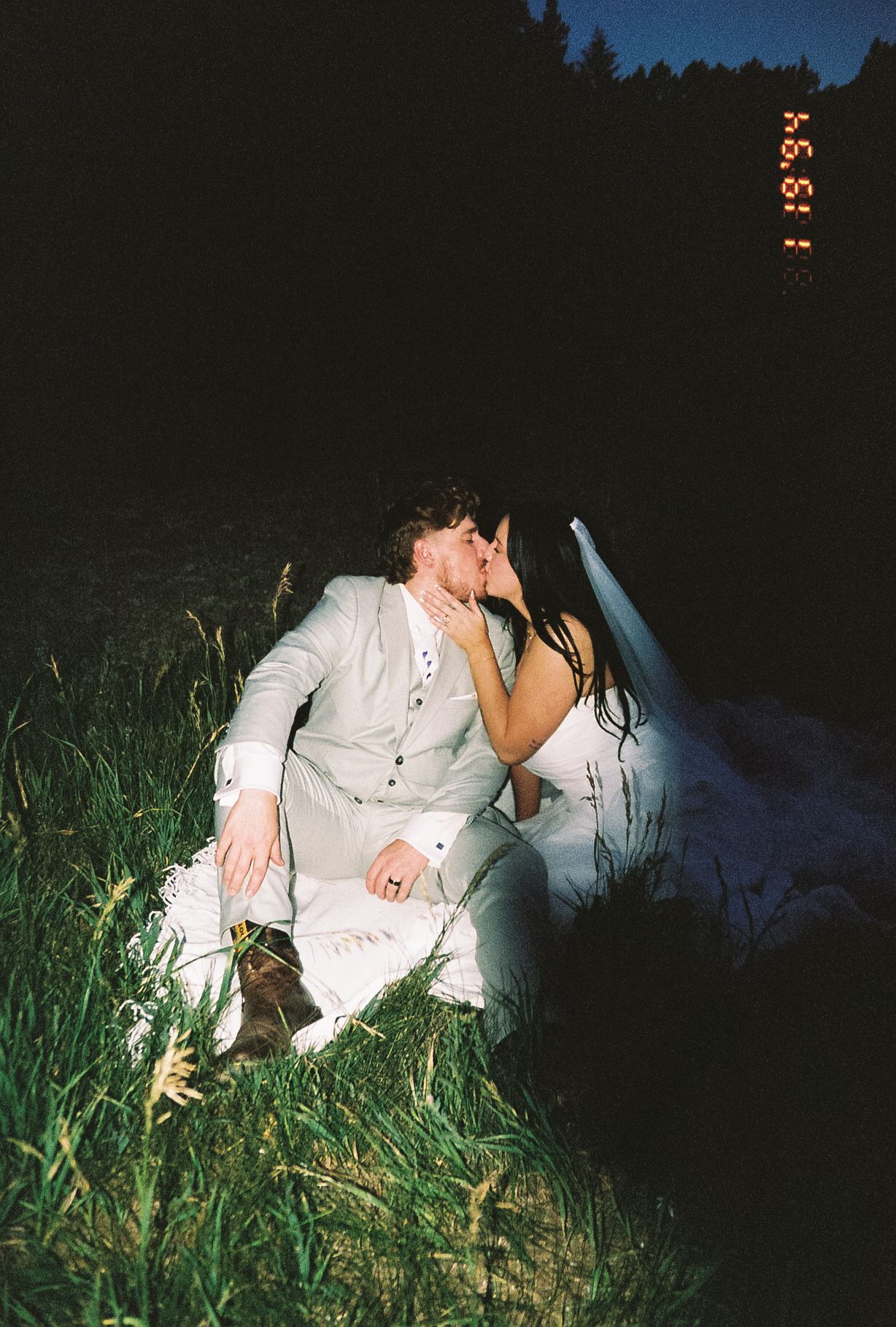 Bride and groom sharing a romantic kiss while seated in tall grass during their nighttime outdoor wedding.