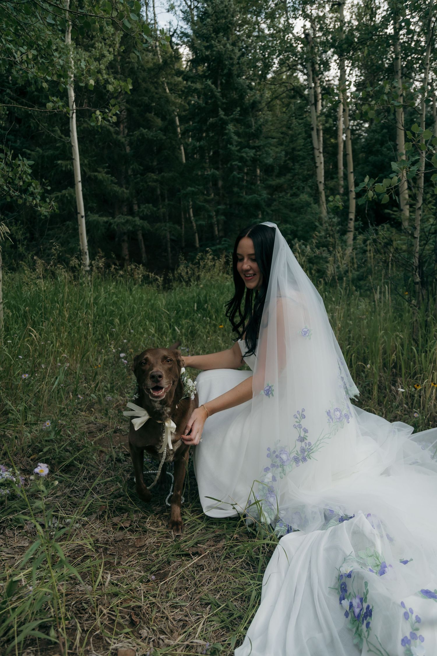 Smiling bride in floral veil kneels beside her chocolate Labrador dog in a green forest clearing.