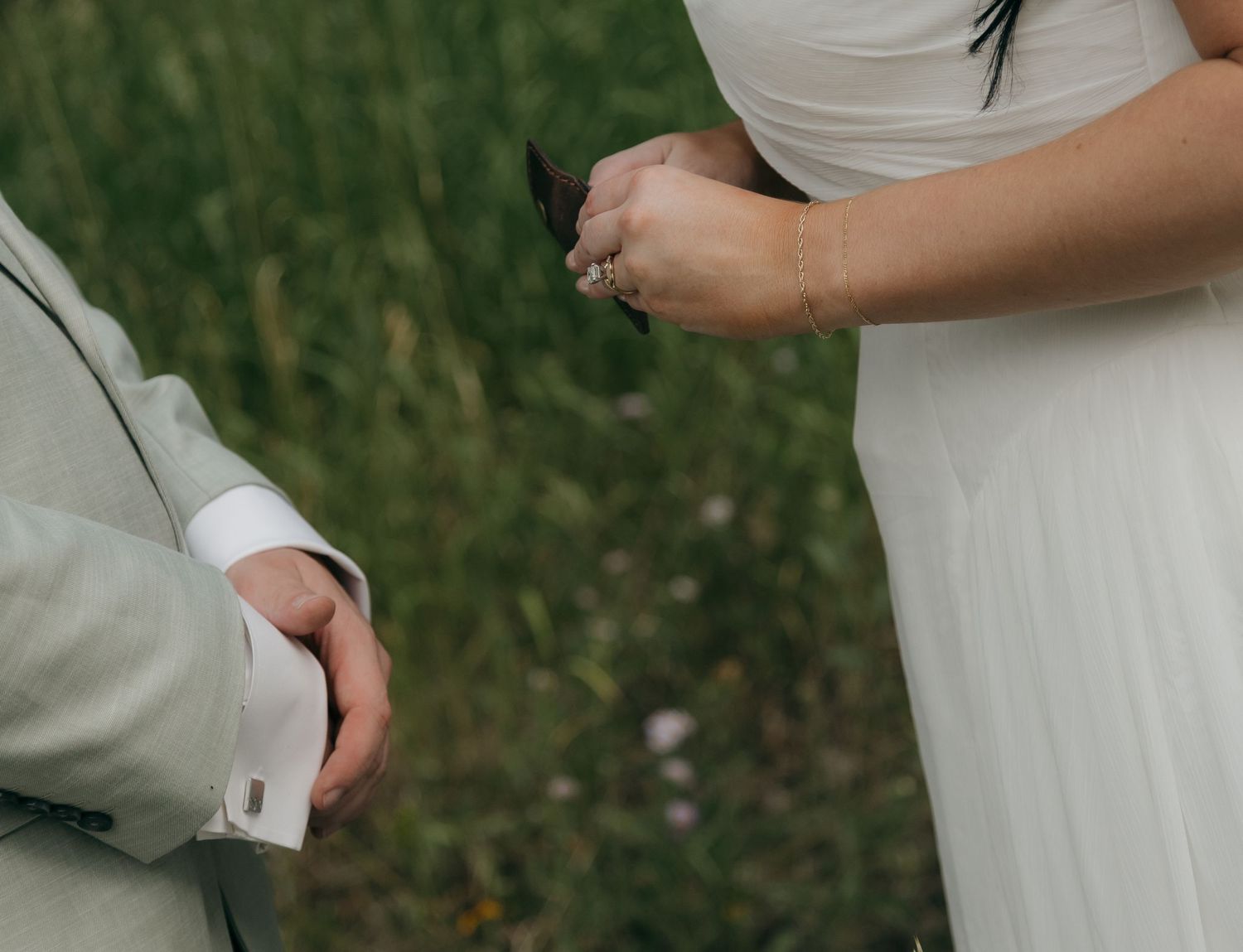 Close-up of bride and groom holding hands during outdoor wedding ceremony in green meadow.