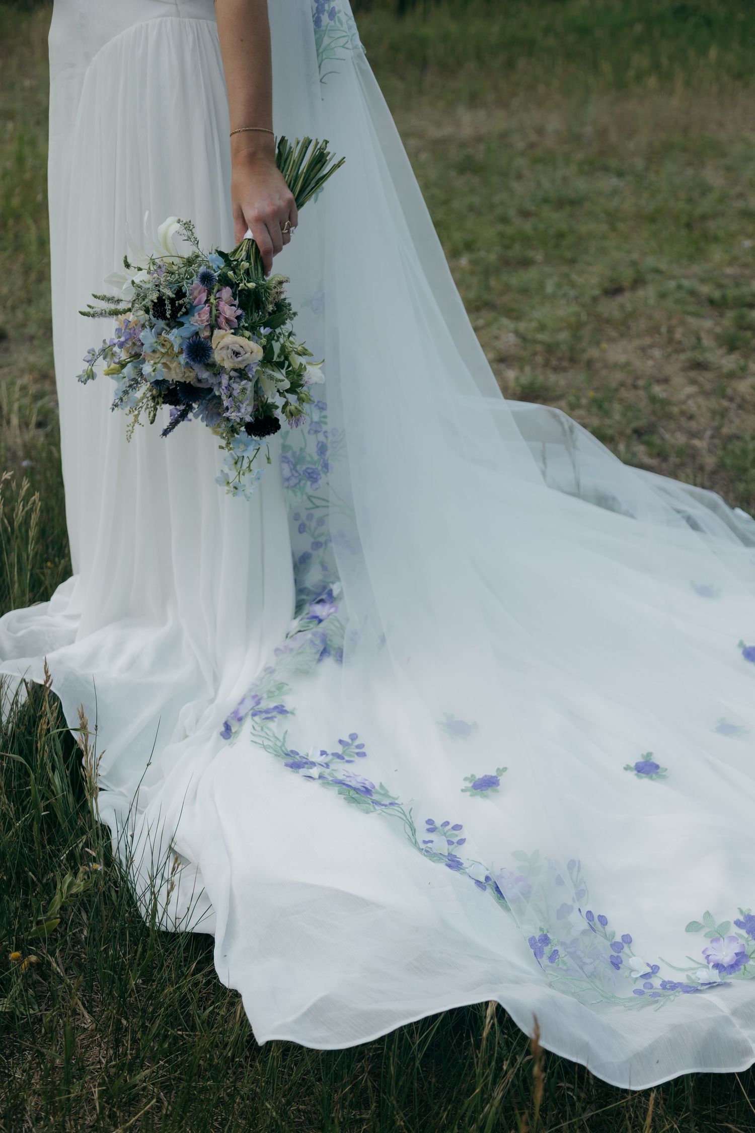 Close-up of a bride holding a blue and white floral bouquet beside her flowing white gown with delicate purple floral embroidery.