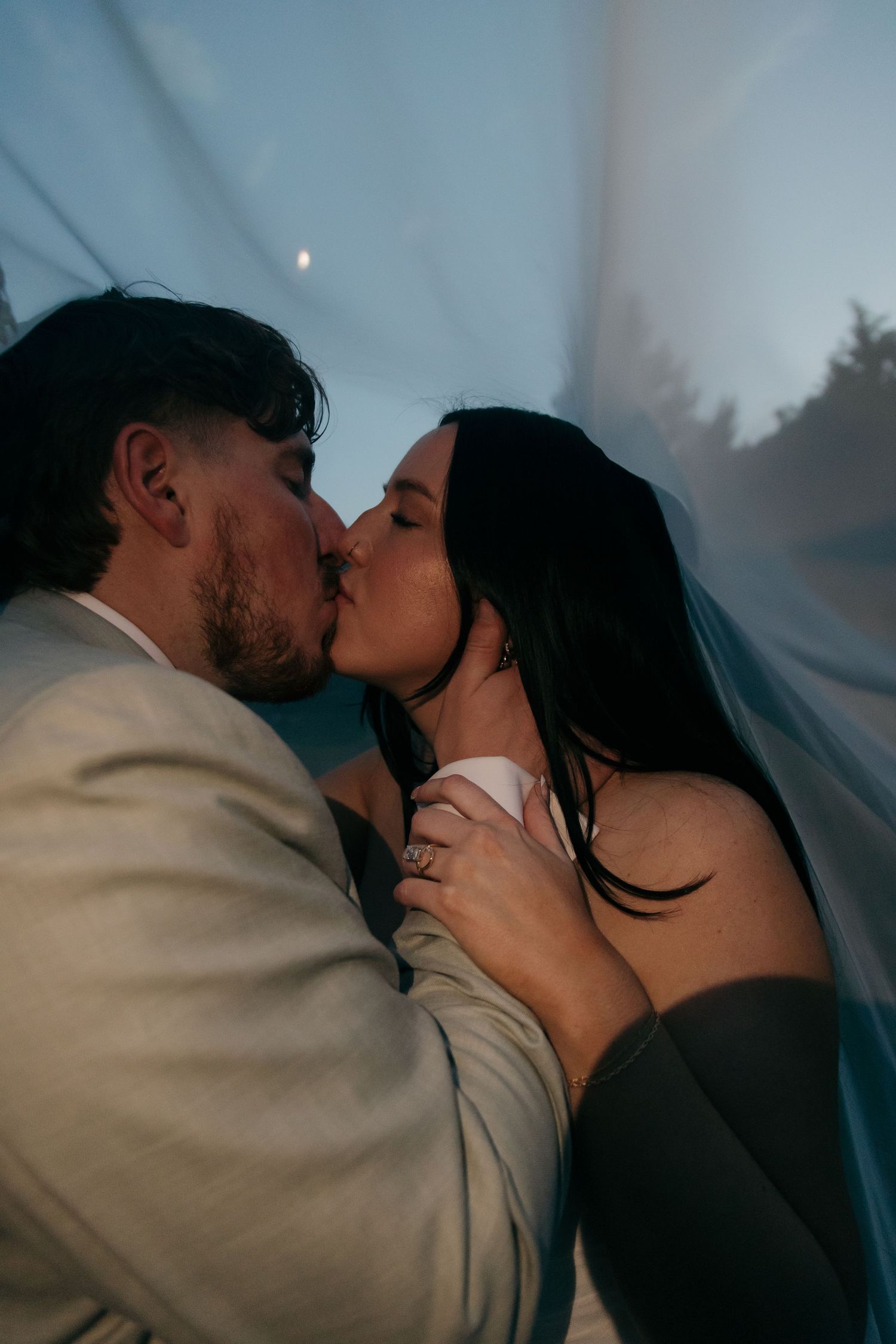 Bride and groom share a passionate kiss beneath a flowing veil in soft blue evening light.