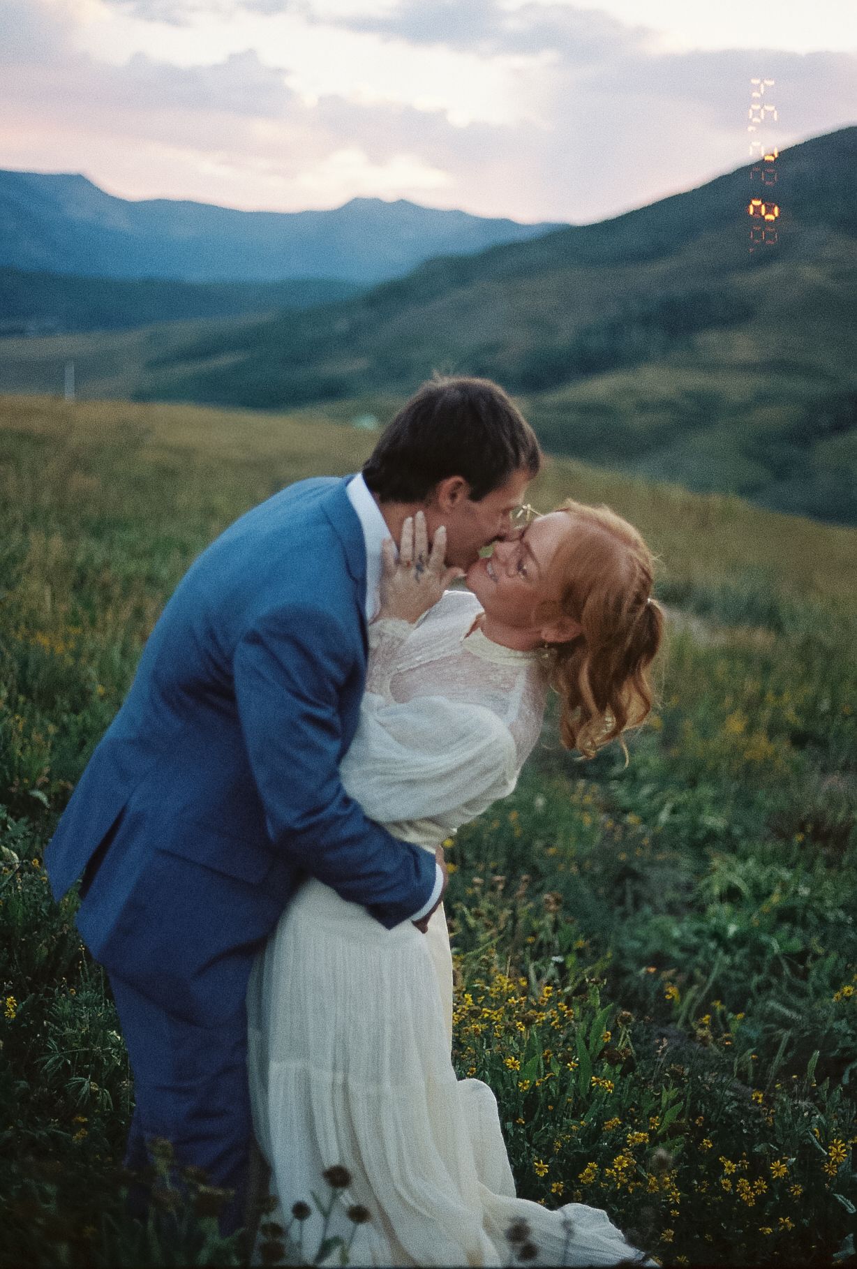 Groom dips bride for romantic kiss in wildflower mountain meadow at sunset during outdoor elopement.