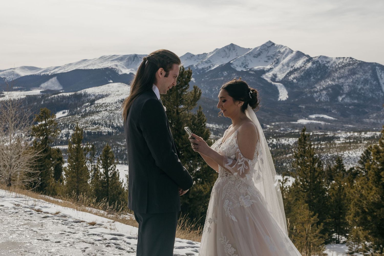 A couple shares a romantic winter wedding moment on a snowy mountain peak with dramatic snow-capped peaks in the background.