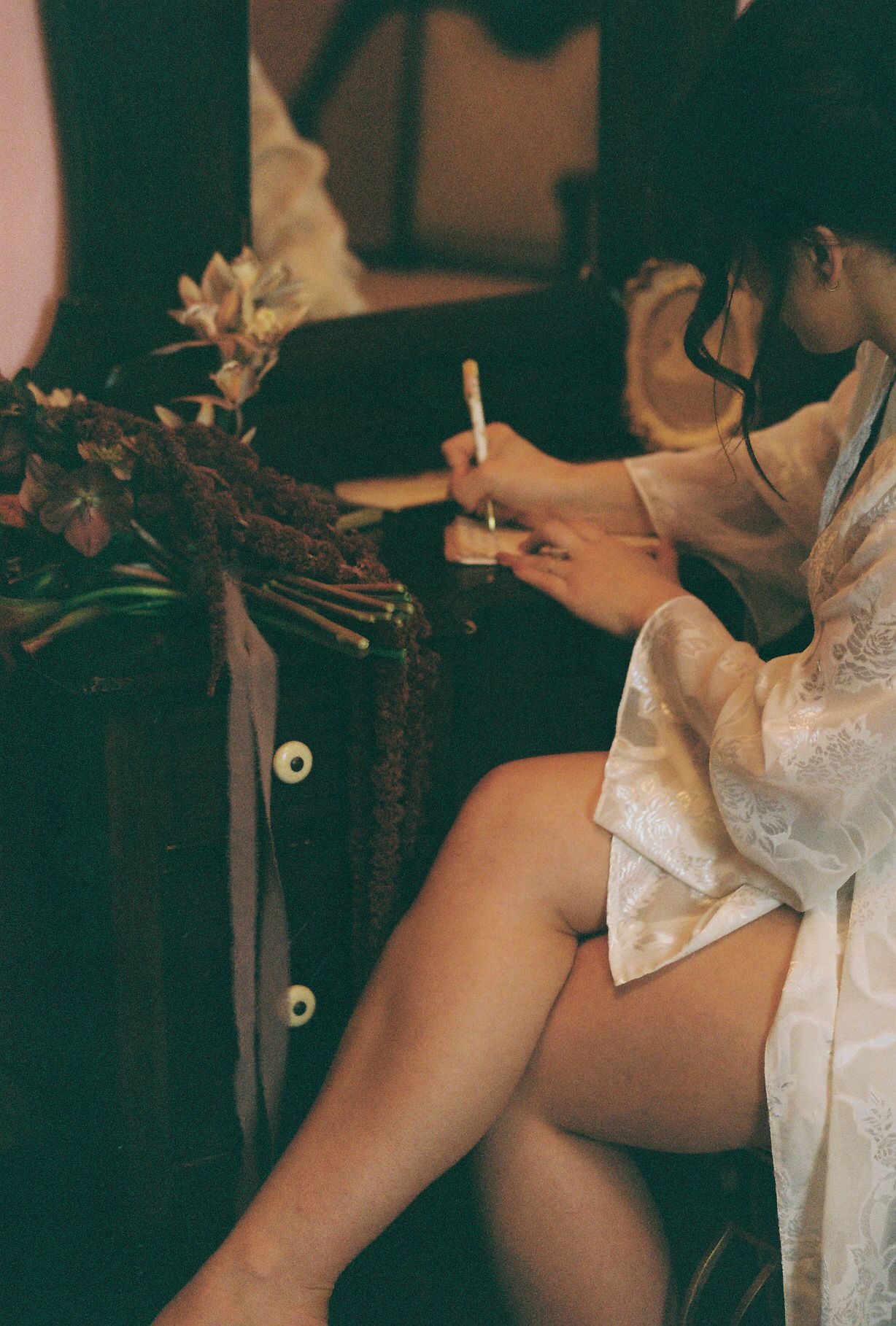 A person in white lace writing while sitting with flowers nearby in vintage-style lighting.