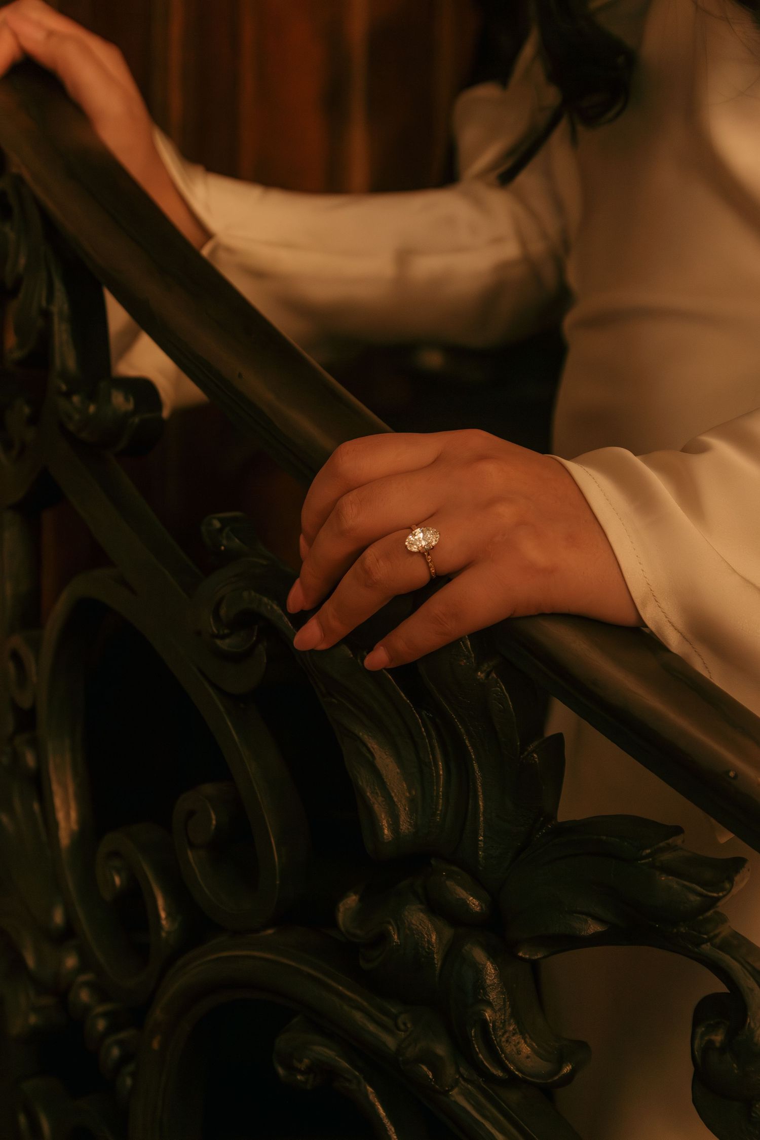 Close-up of a hand wearing an engagement ring resting on an ornate dark railing in warm lighting.