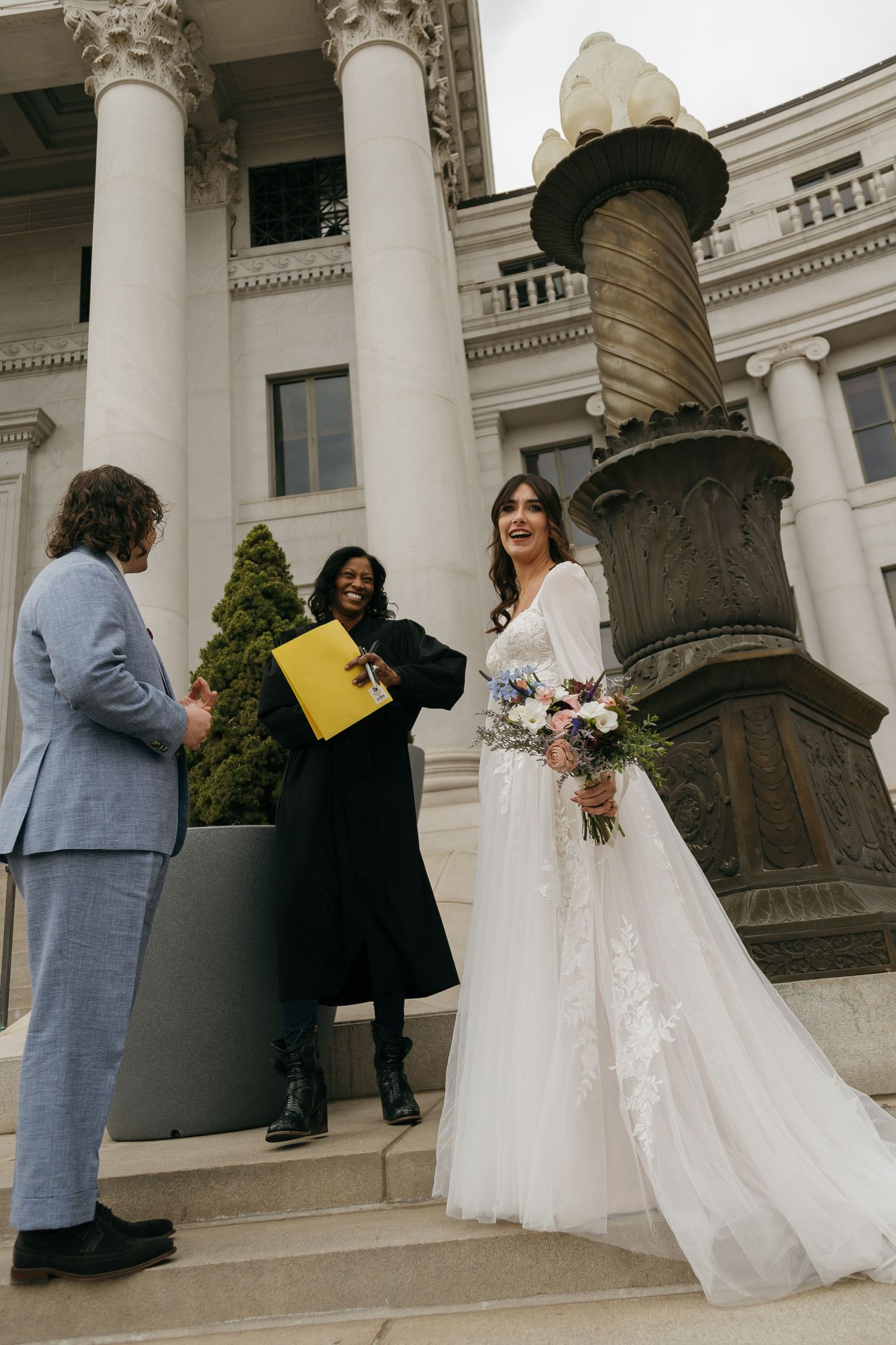 A wedding party gathers on grand stone steps of a neoclassical building with white columns.