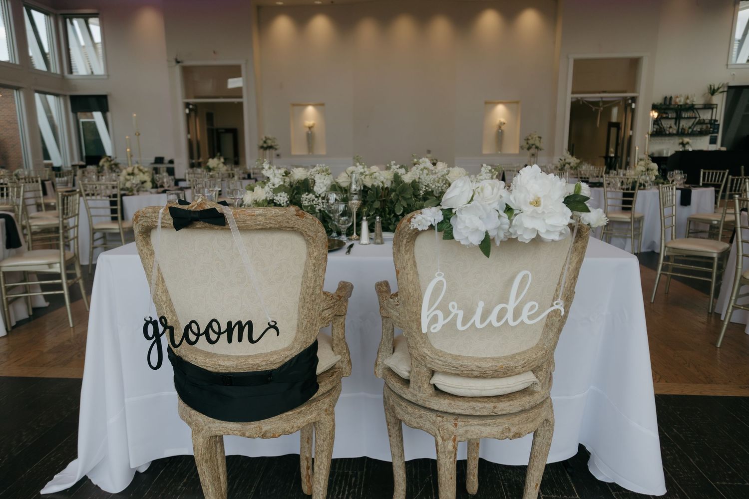 Elegant wedding reception sweetheart table with groom and bride chairs, white florals, and gold decor.
