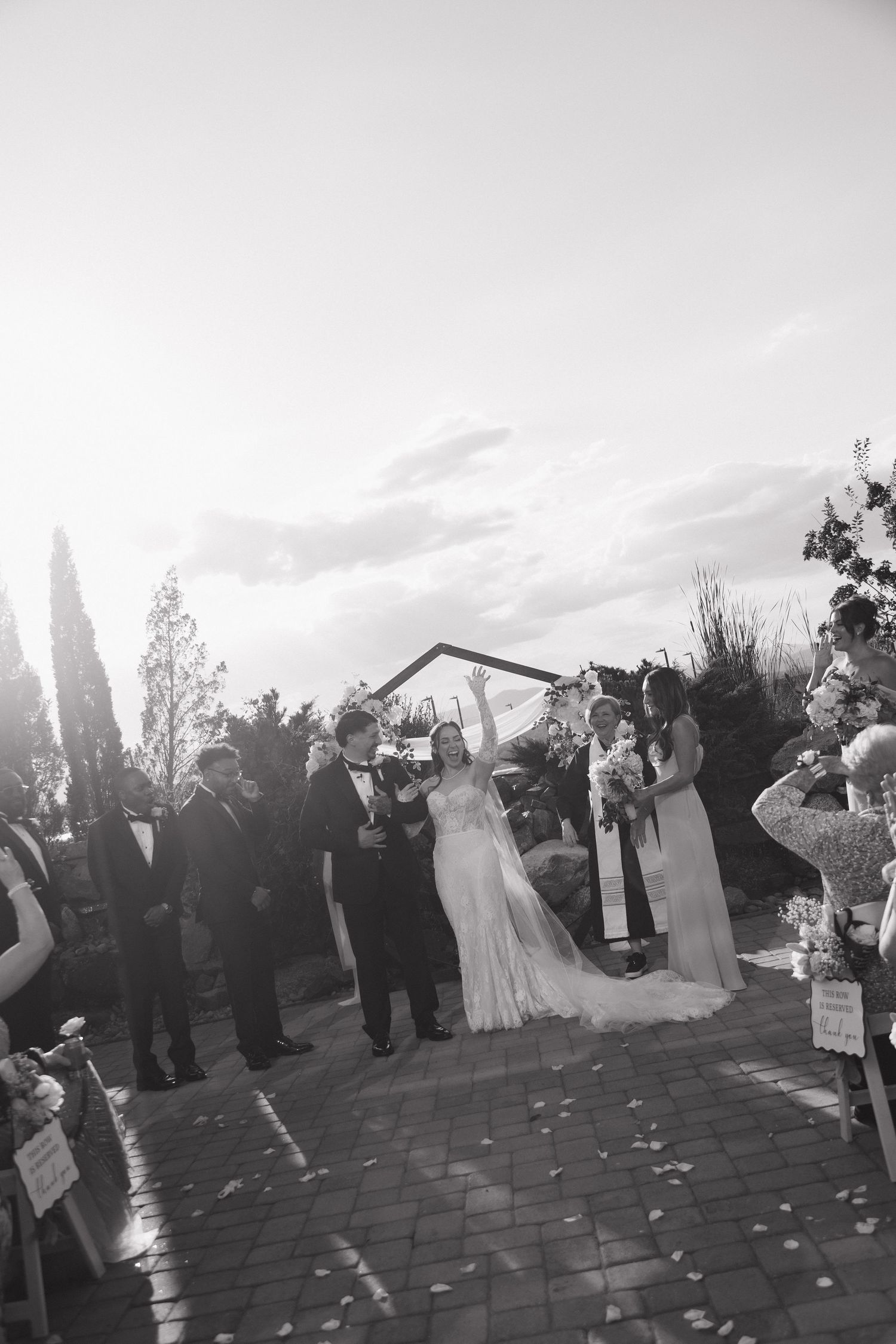 Black and white photo of newlyweds celebrating at outdoor wedding ceremony with guests cheering.