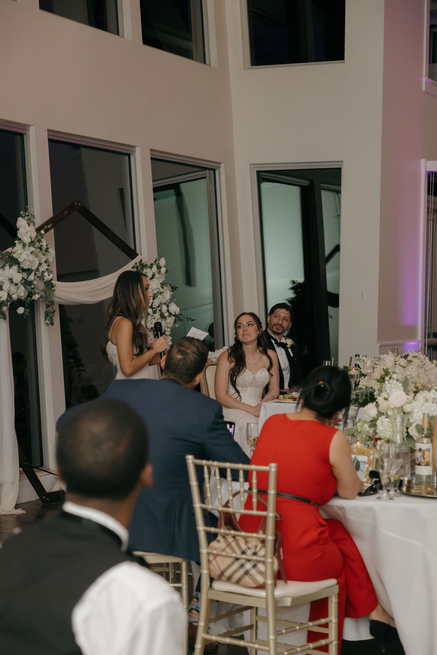 Wedding guest delivers speech to smiling bride and groom at elegant indoor reception dinner.