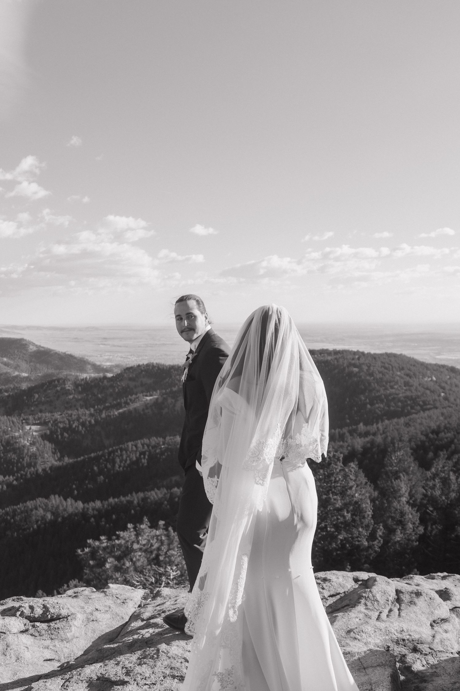 A wedding couple stands on a mountain overlook with sweeping views of forested hills under a bright sky.