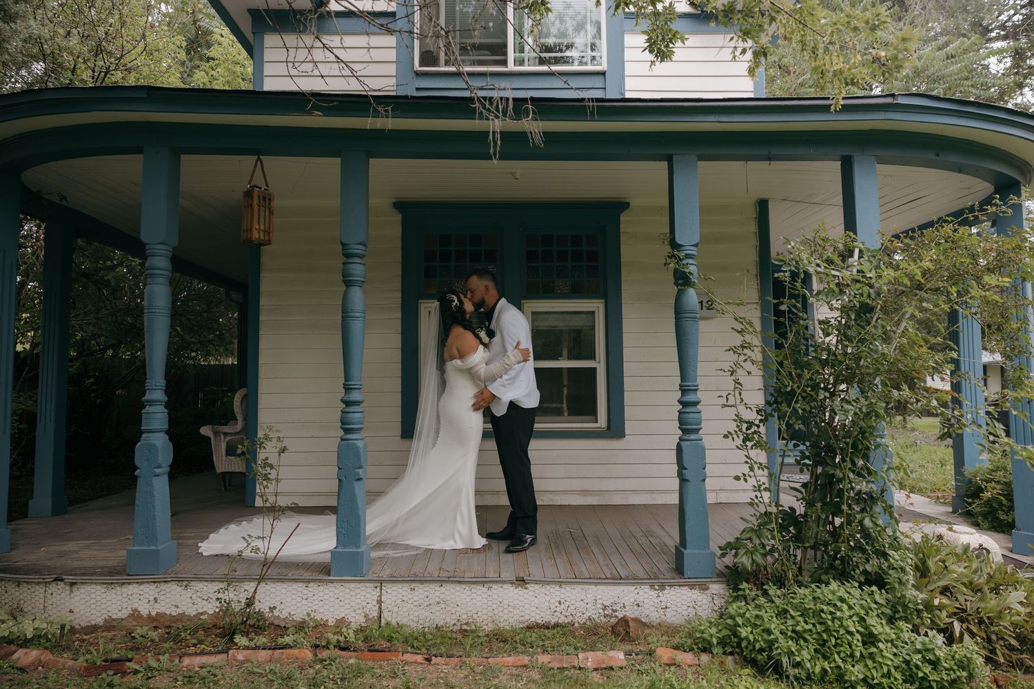 Bride and groom sharing a kiss on a charming blue-columned porch of a white Victorian house on their wedding day.