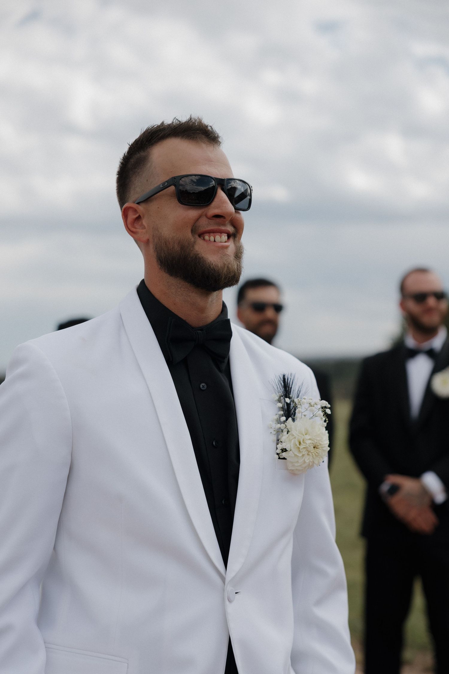 Smiling groom in white tuxedo jacket with boutonniere and sunglasses at outdoor wedding ceremony.
