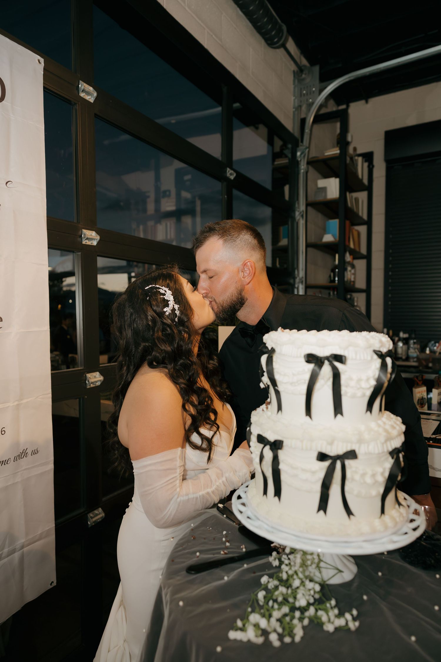 Bride and groom kissing beside white wedding cake with black bow decorations.