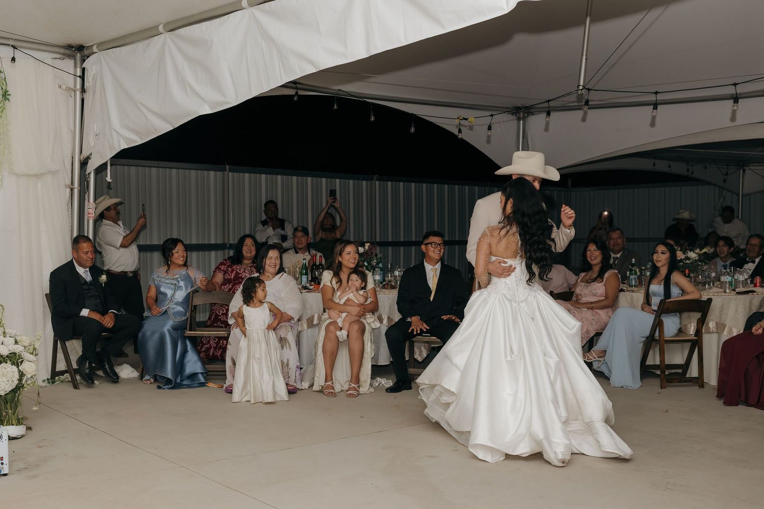 Bride and groom share first dance under white tent while wedding guests watch at elegant outdoor reception.