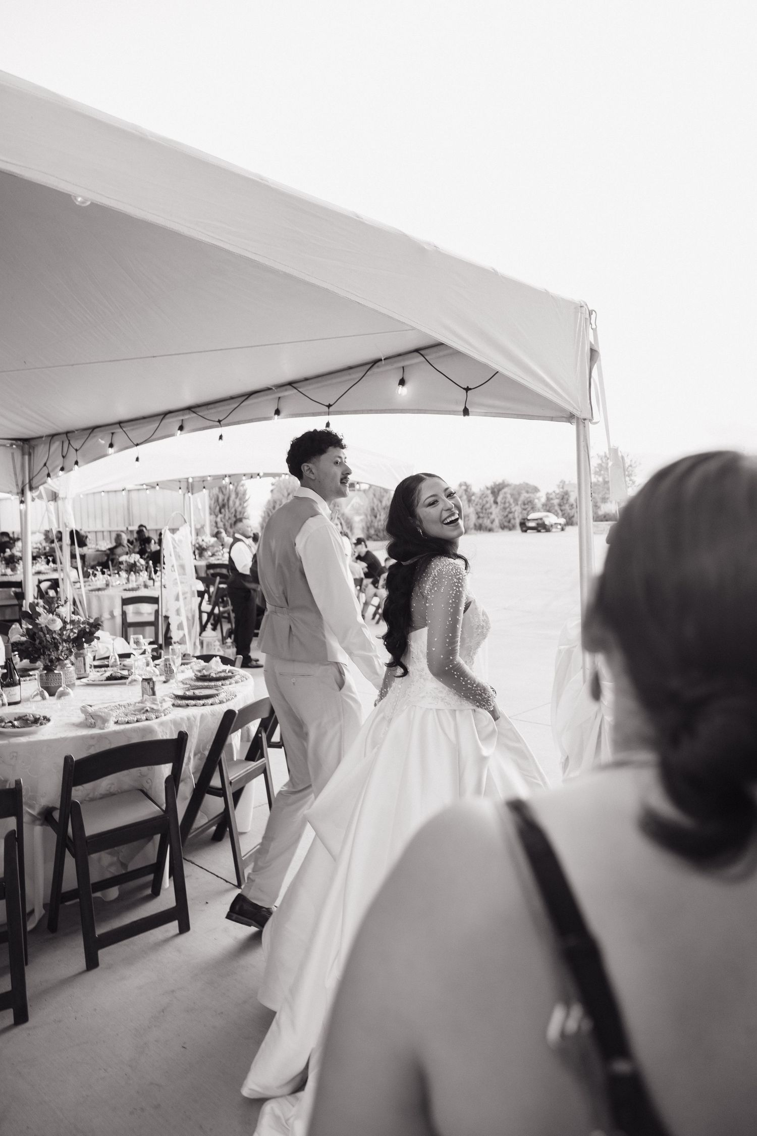 Black and white photo of laughing bride and groom making grand entrance under white tent at outdoor wedding reception.