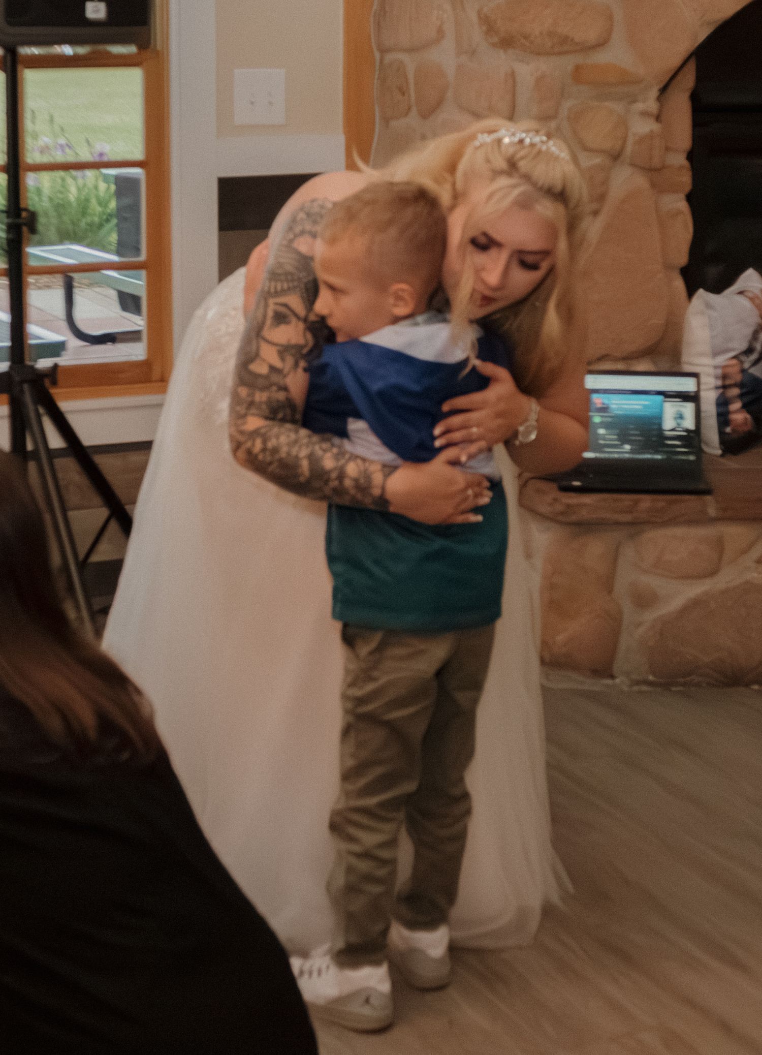 A heartwarming embrace between a young child and a bride in a white dress with sleeve tattoos at a wedding reception.