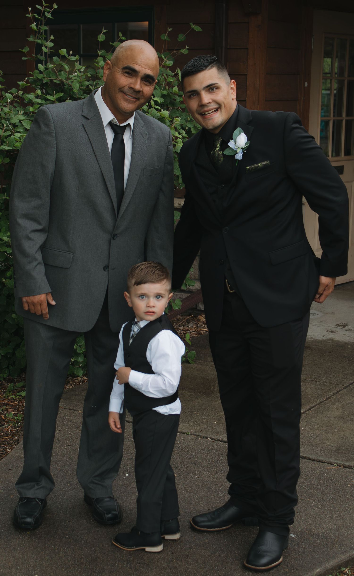 Three generations pose together in formal attire outside a rustic venue.