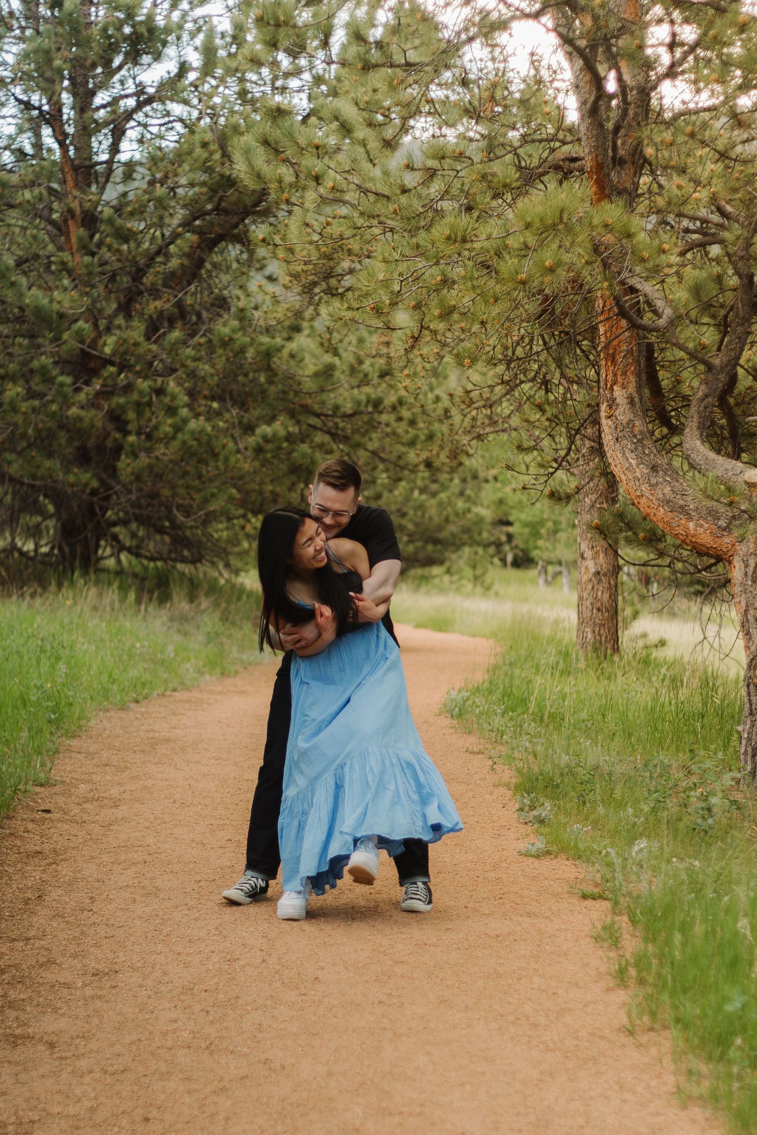 A couple dances together on a dirt path surrounded by tall pine trees.