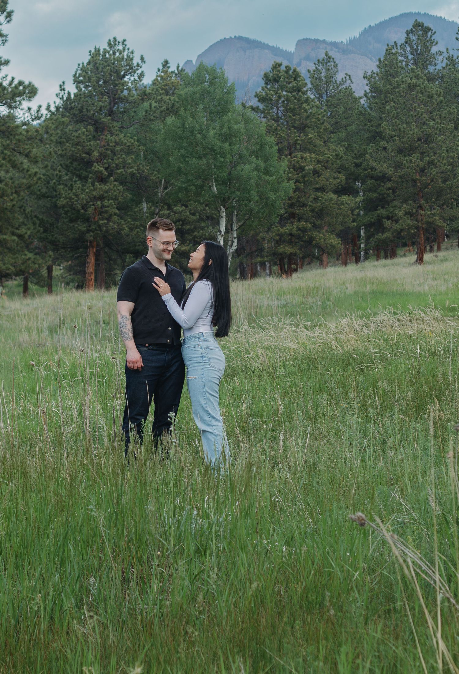 A couple stands together in a mountain meadow with evergreen trees and mountains in the background.