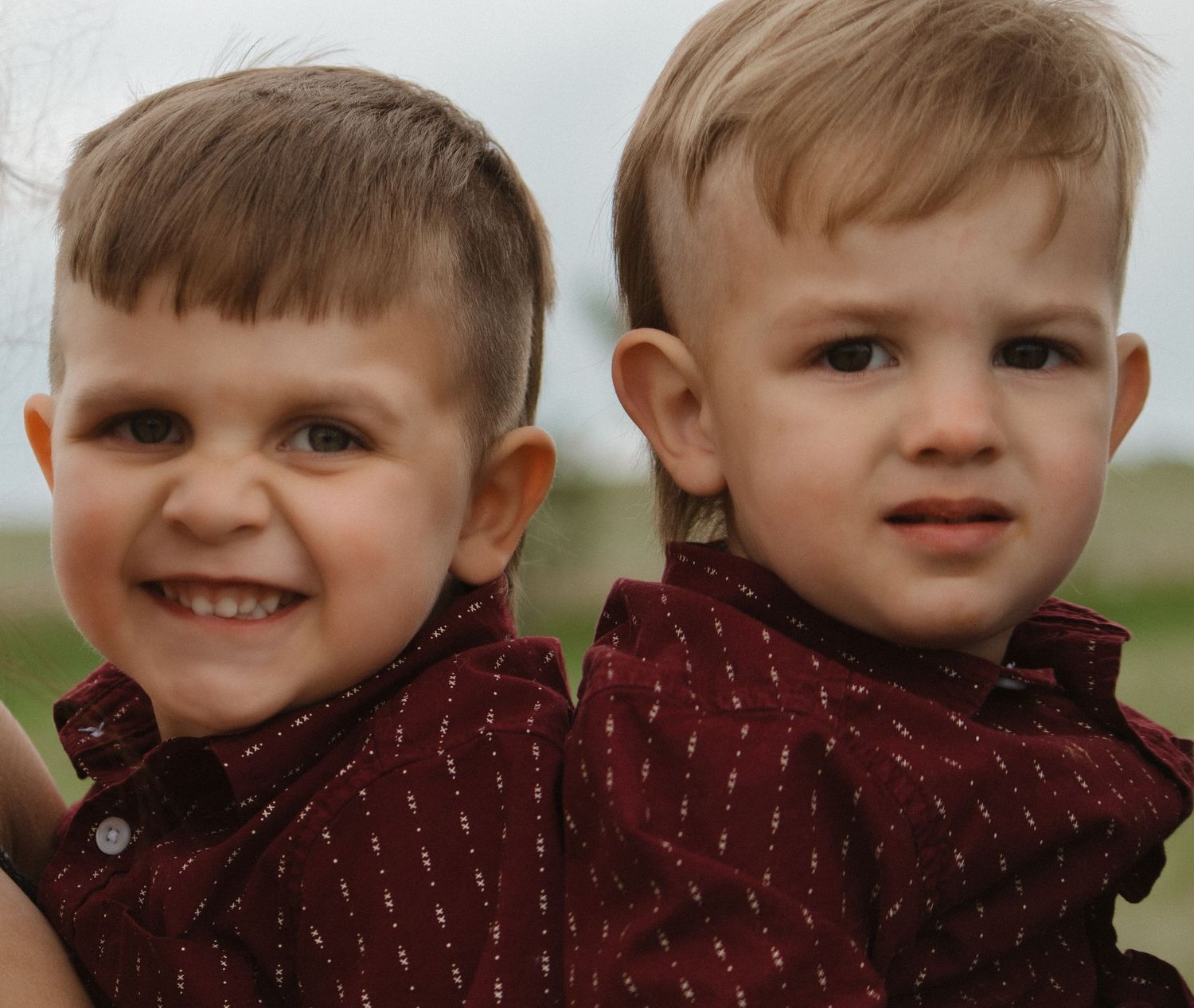 Two young children wearing matching burgundy shirts smile at the camera outdoors.