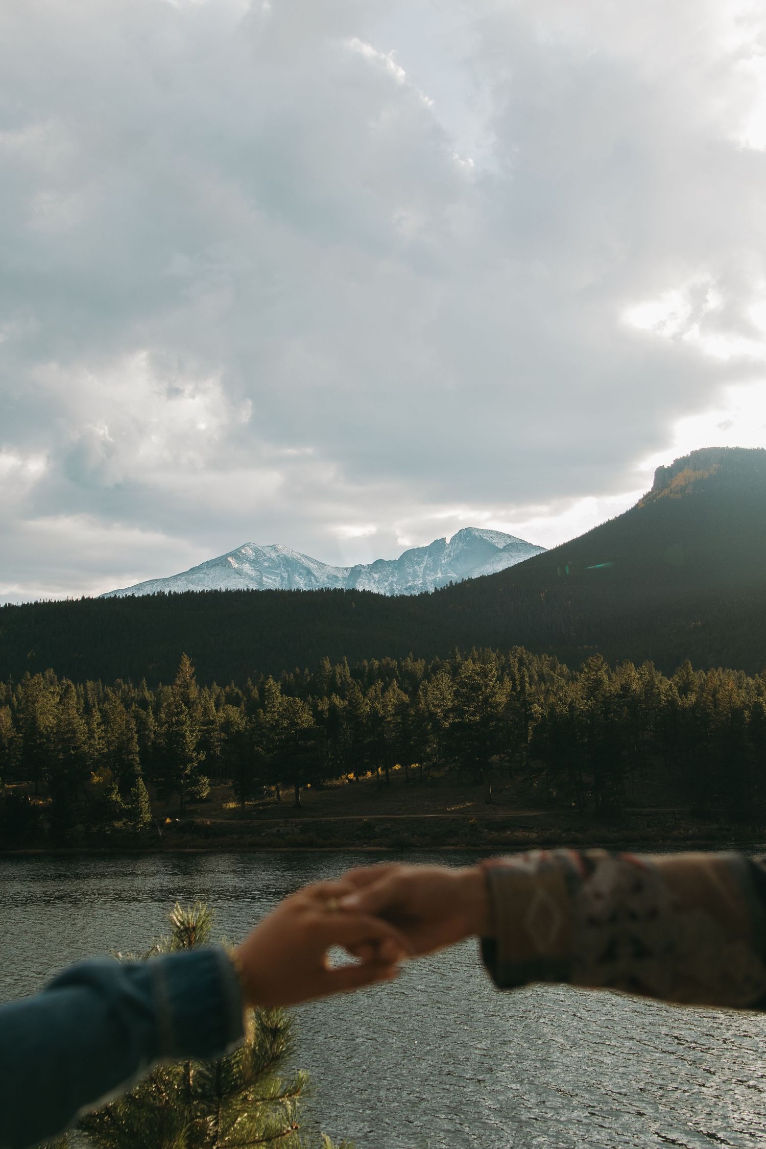 Snow-capped mountains rise above a serene lake surrounded by evergreen forest under moody cloudy skies.