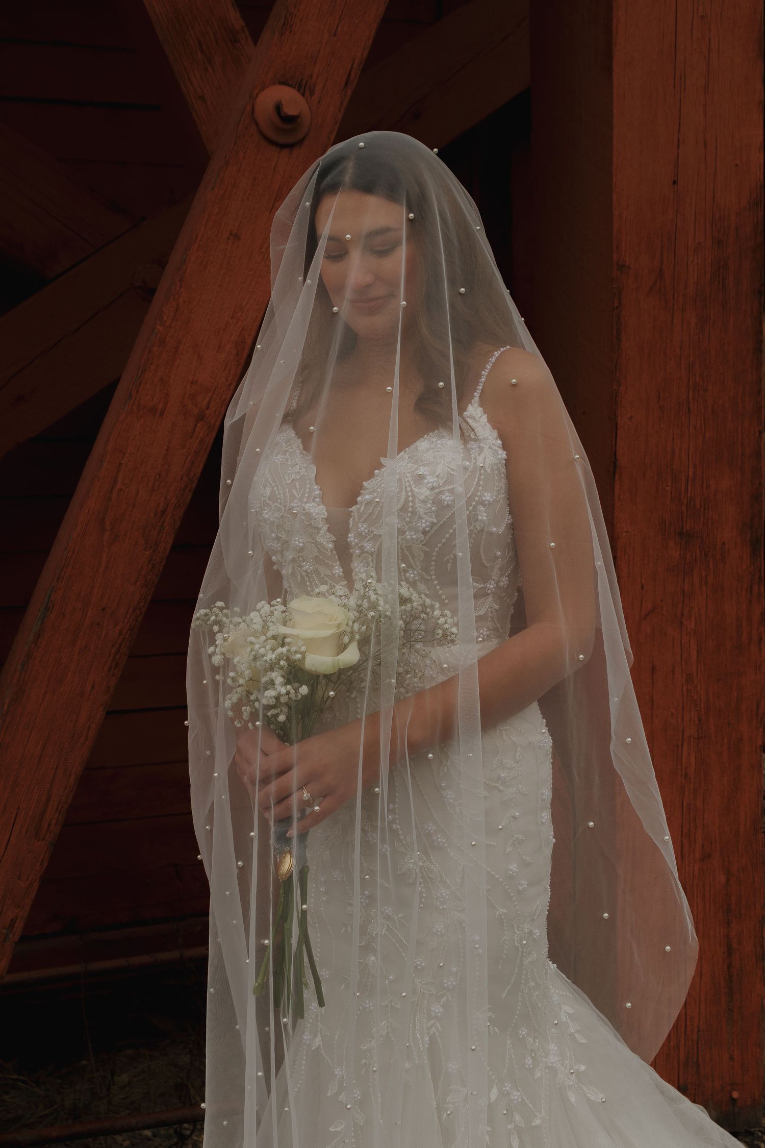 A bride in a lace wedding gown holds a single rose while wearing a pearl-studded veil.