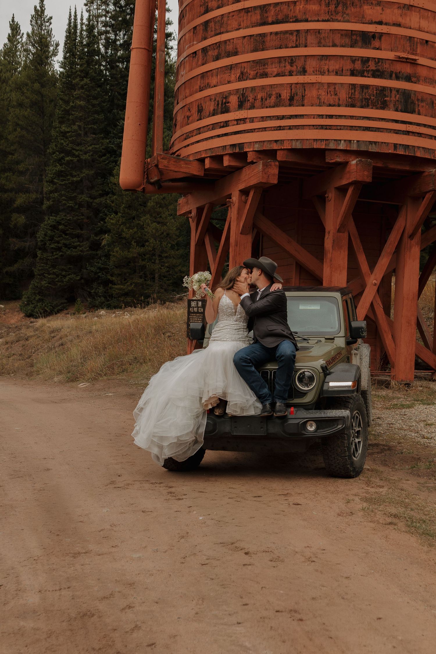 A couple poses near a rustic wooden water tower structure in a mountain setting.