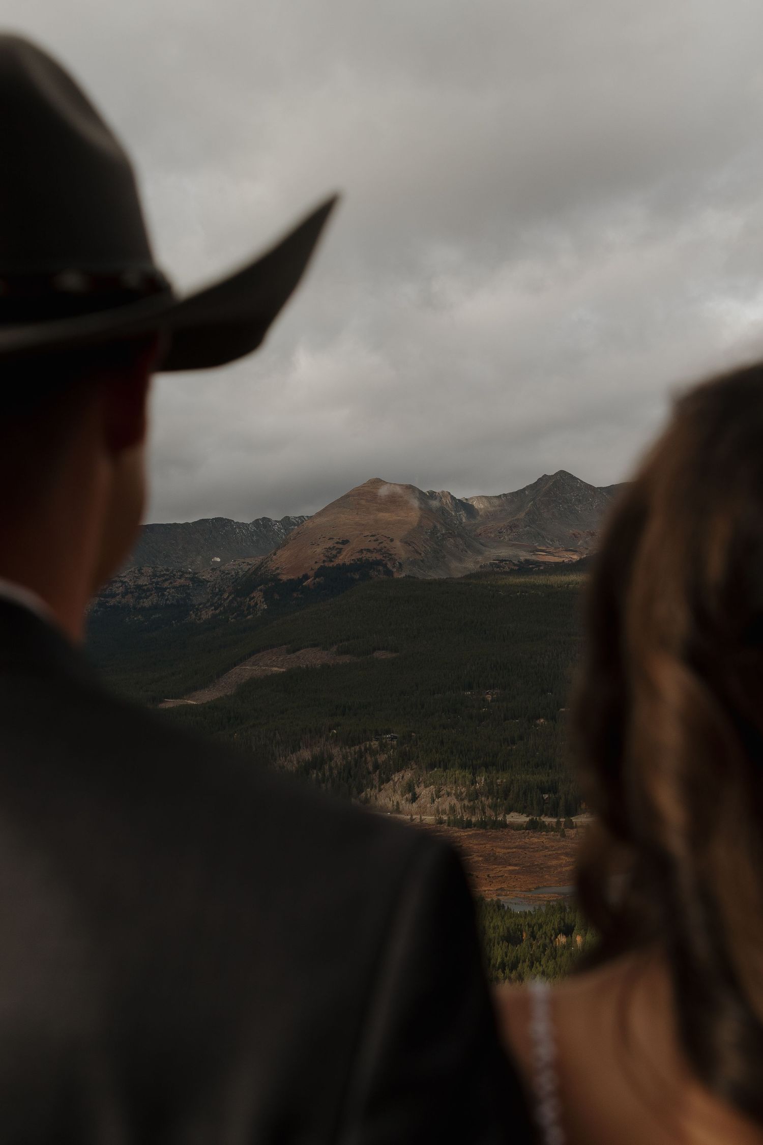 A scenic mountain vista captured from behind two silhouetted figures wearing western attire.