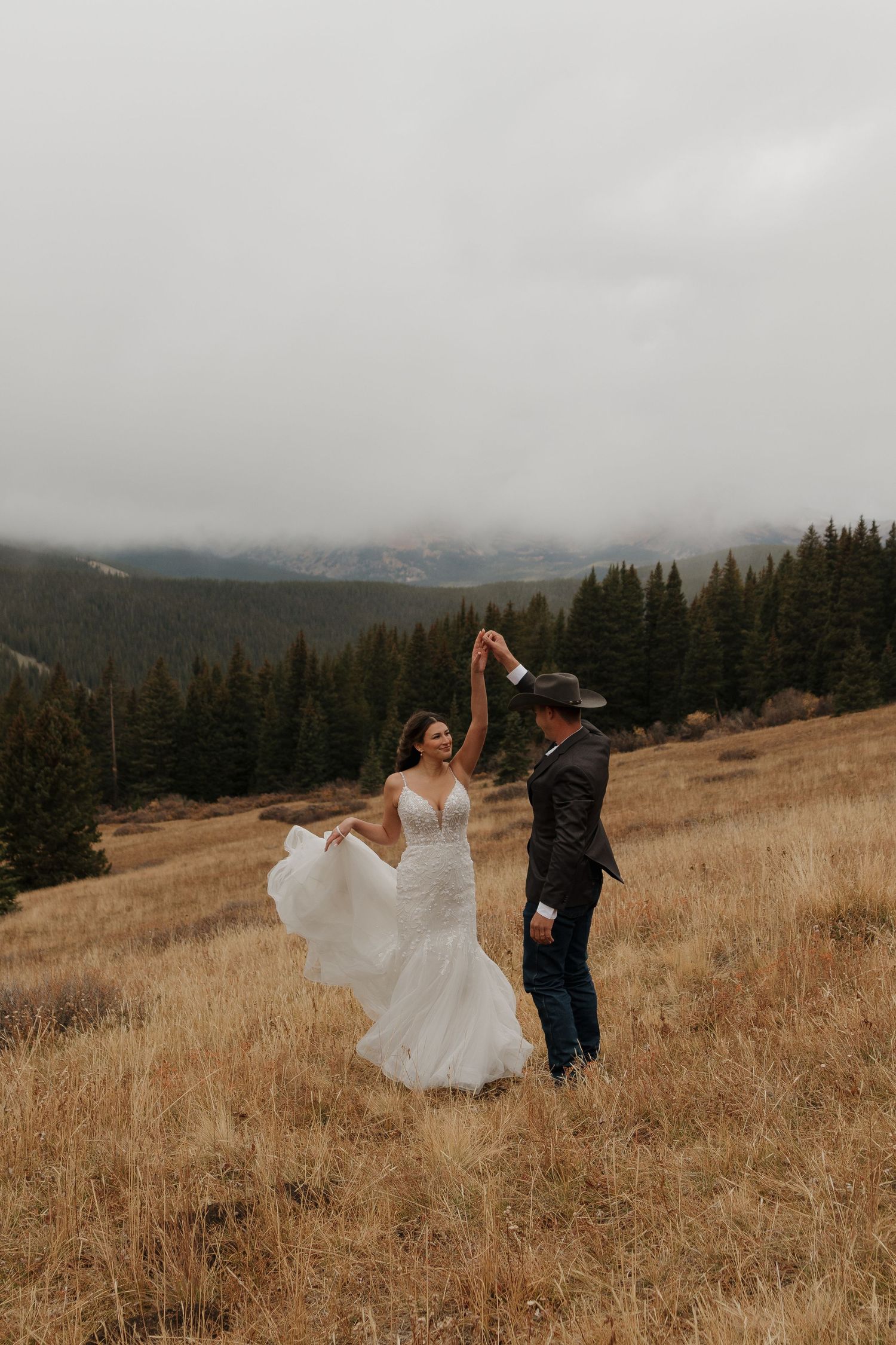 A couple dances together on a mountainside meadow with evergreen forests in the background.