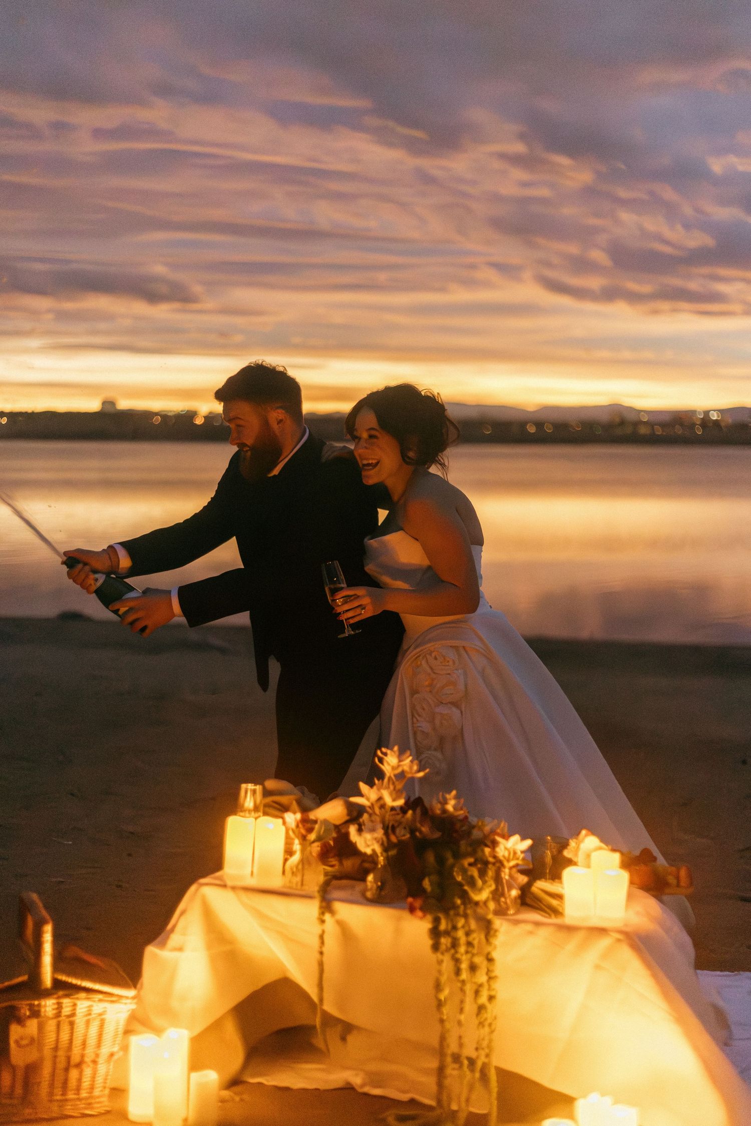 Romantic sunset wedding scene with couple toasting by candlelit table near a tranquil lake.