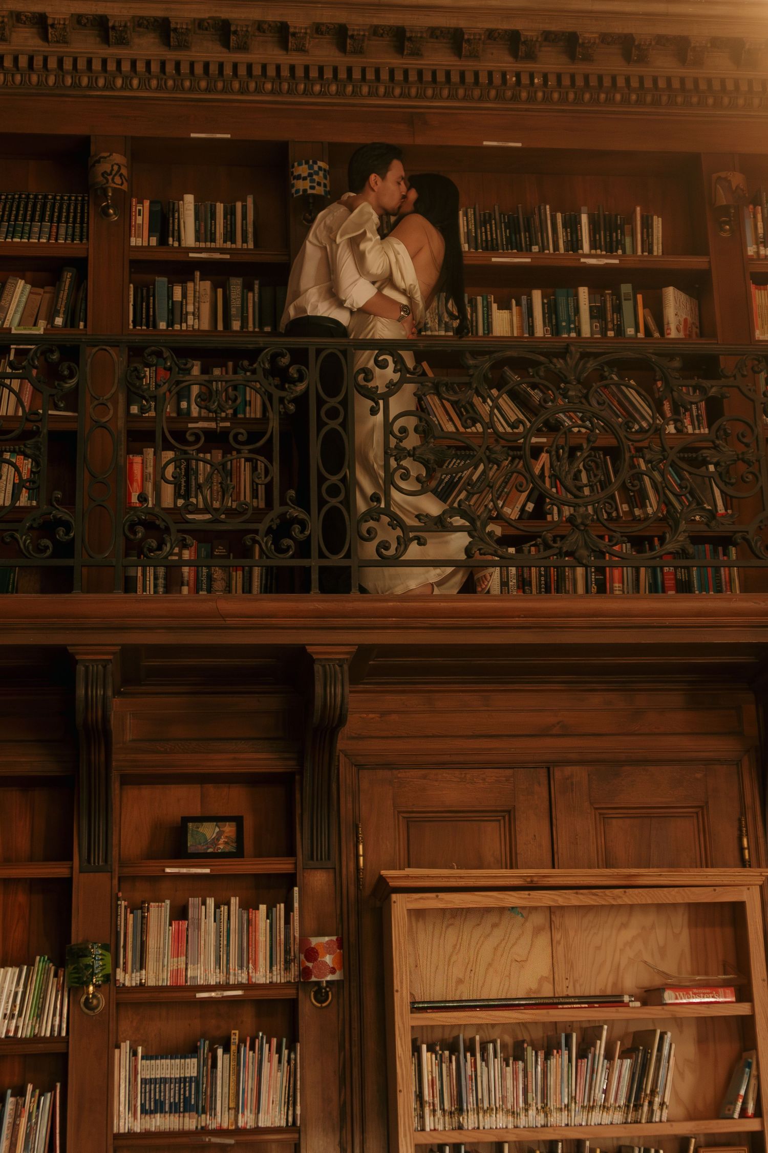 A romantic moment between a couple embracing on a vintage library balcony surrounded by wooden bookshelves.