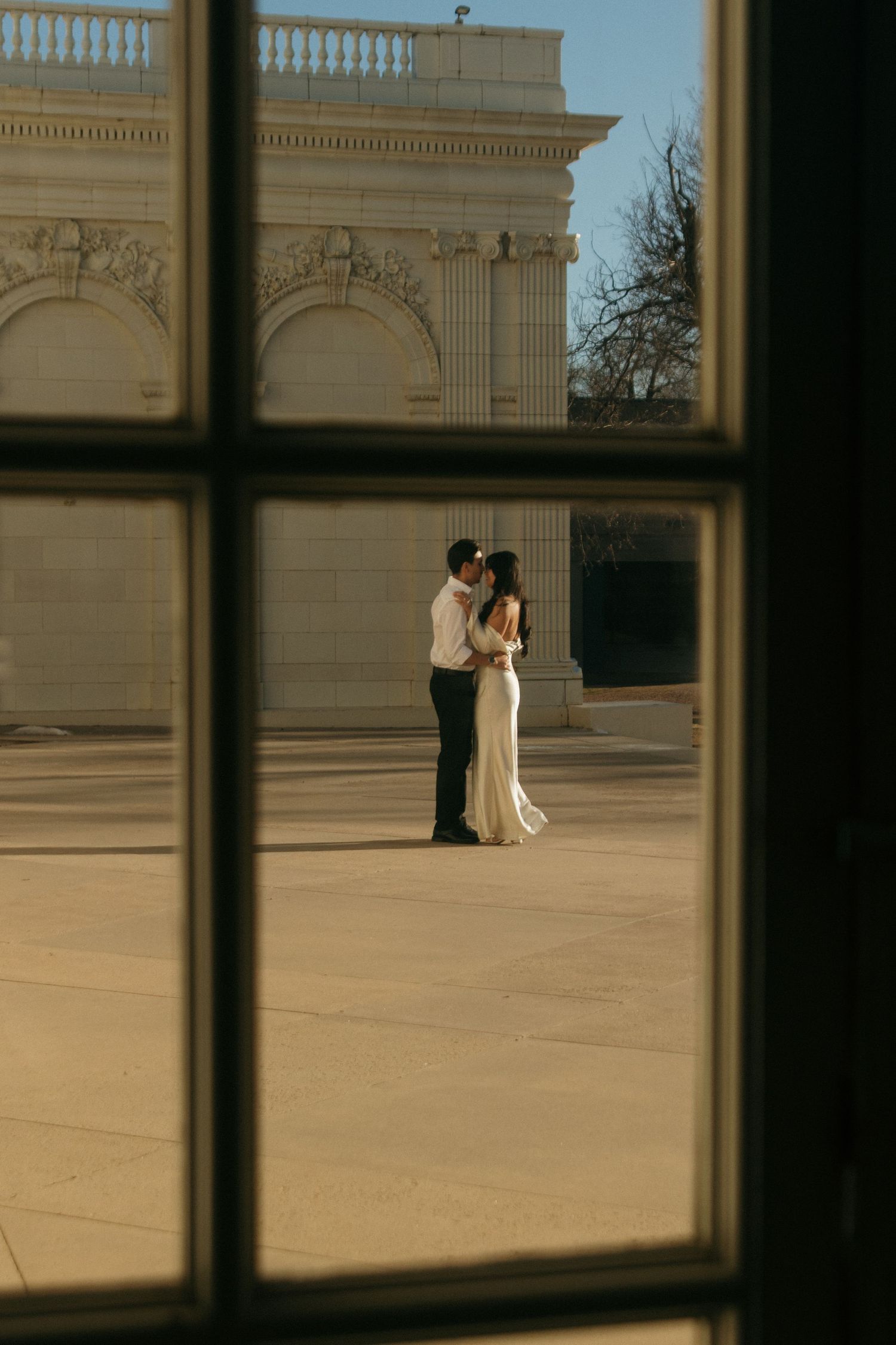 Architectural view through window panes of couple in formal wear on sunlit stone terrace.