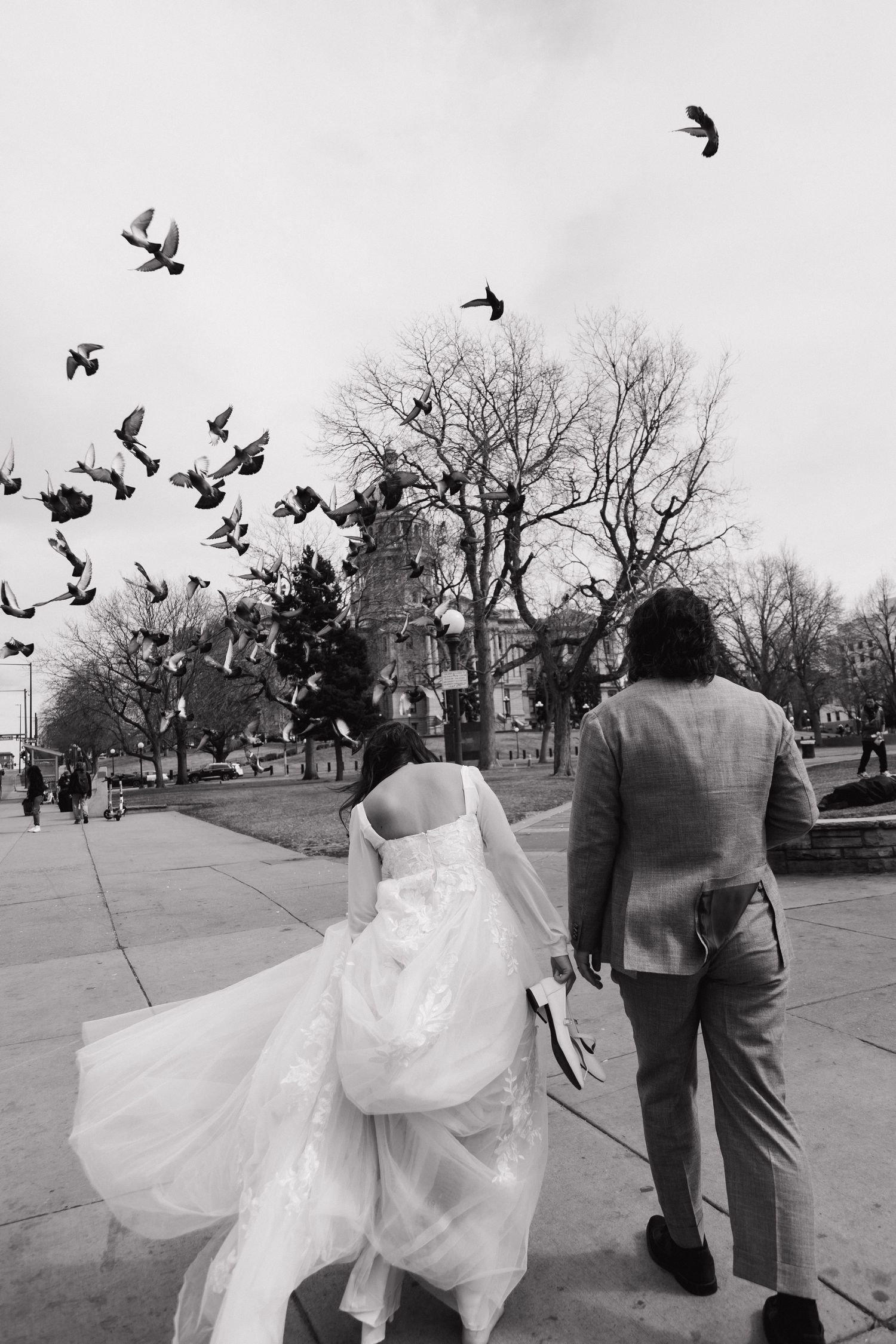 A romantic black and white photo of newlyweds walking as birds fly overhead near a historic building.