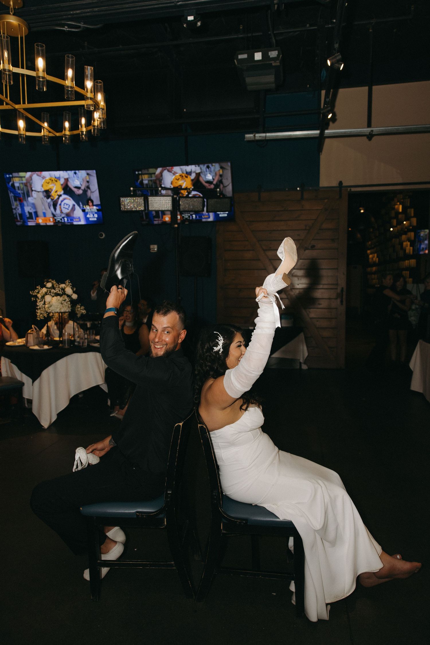 Bride and groom play shoe game at wedding reception, laughing and holding shoes up in the air.