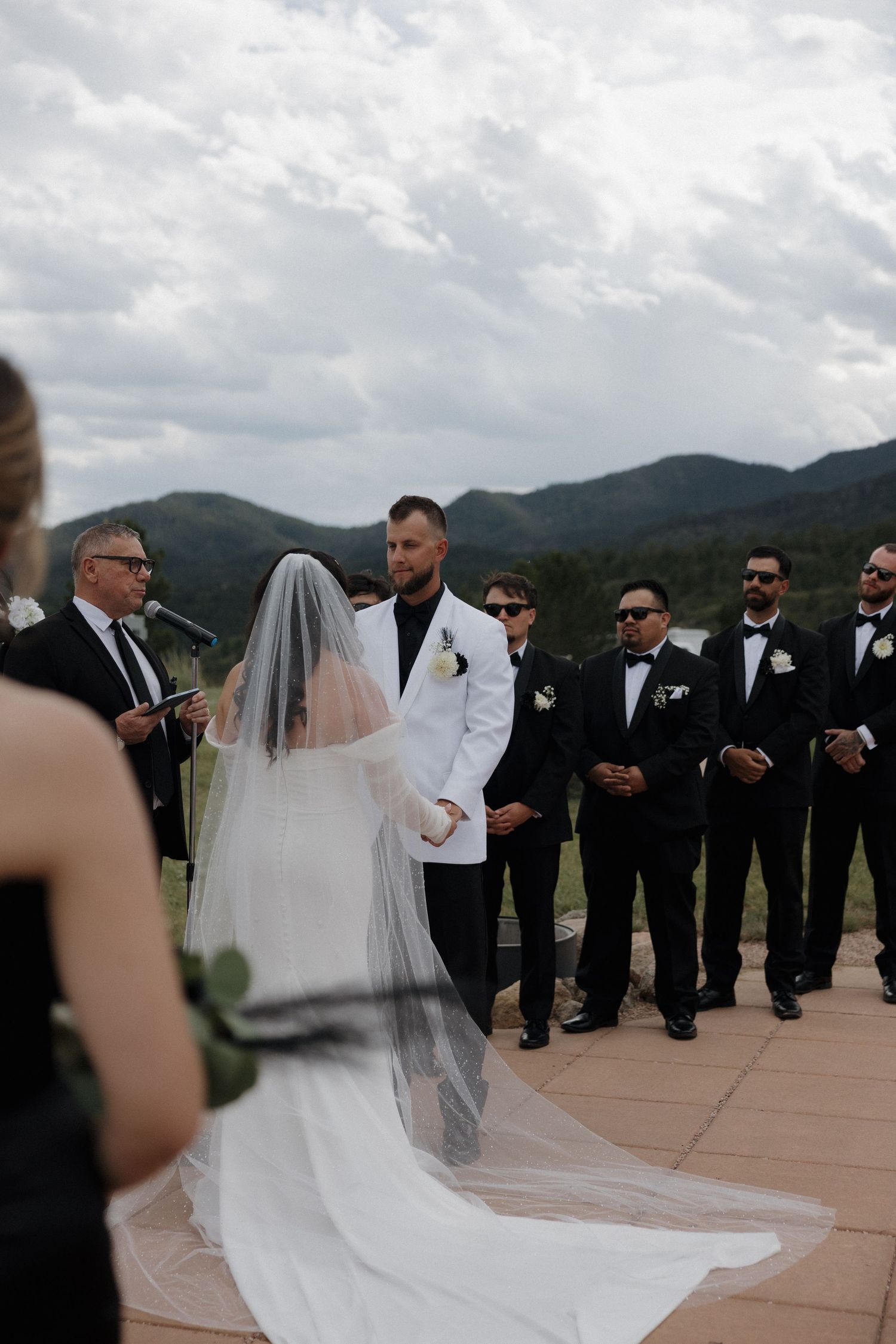 Outdoor mountain wedding ceremony with bride in veil and groom in white jacket.
