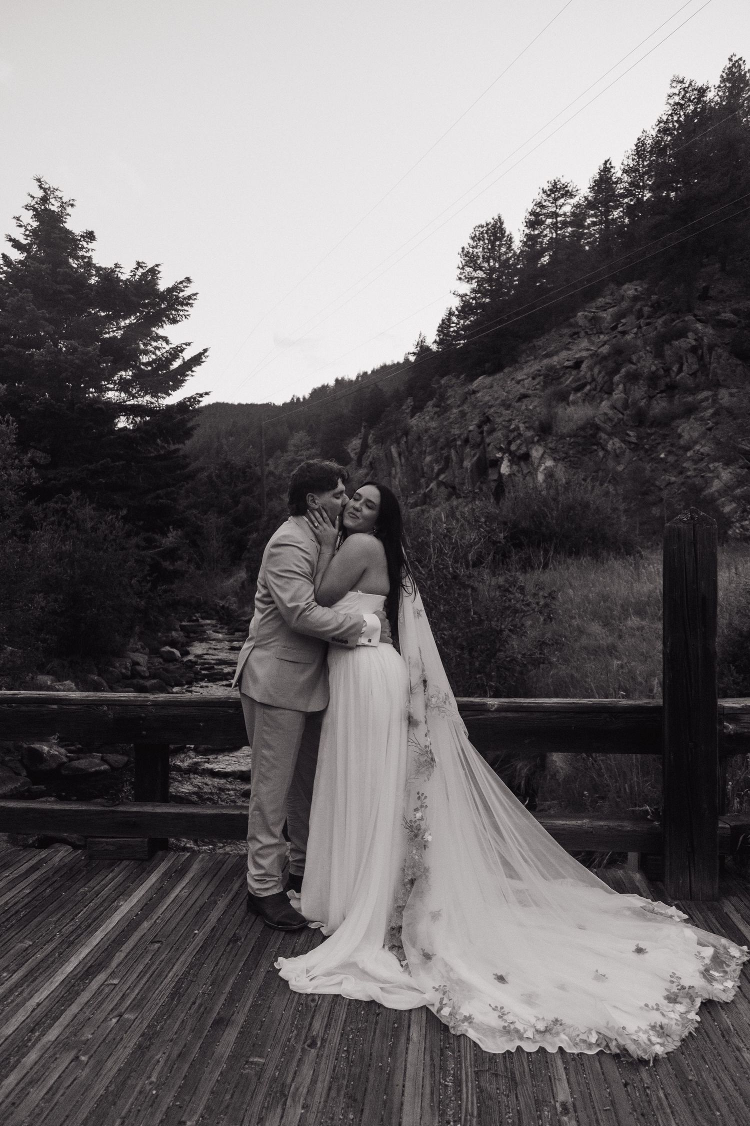 Black and white photo of bride and groom embracing on a wooden bridge surrounded by forest.