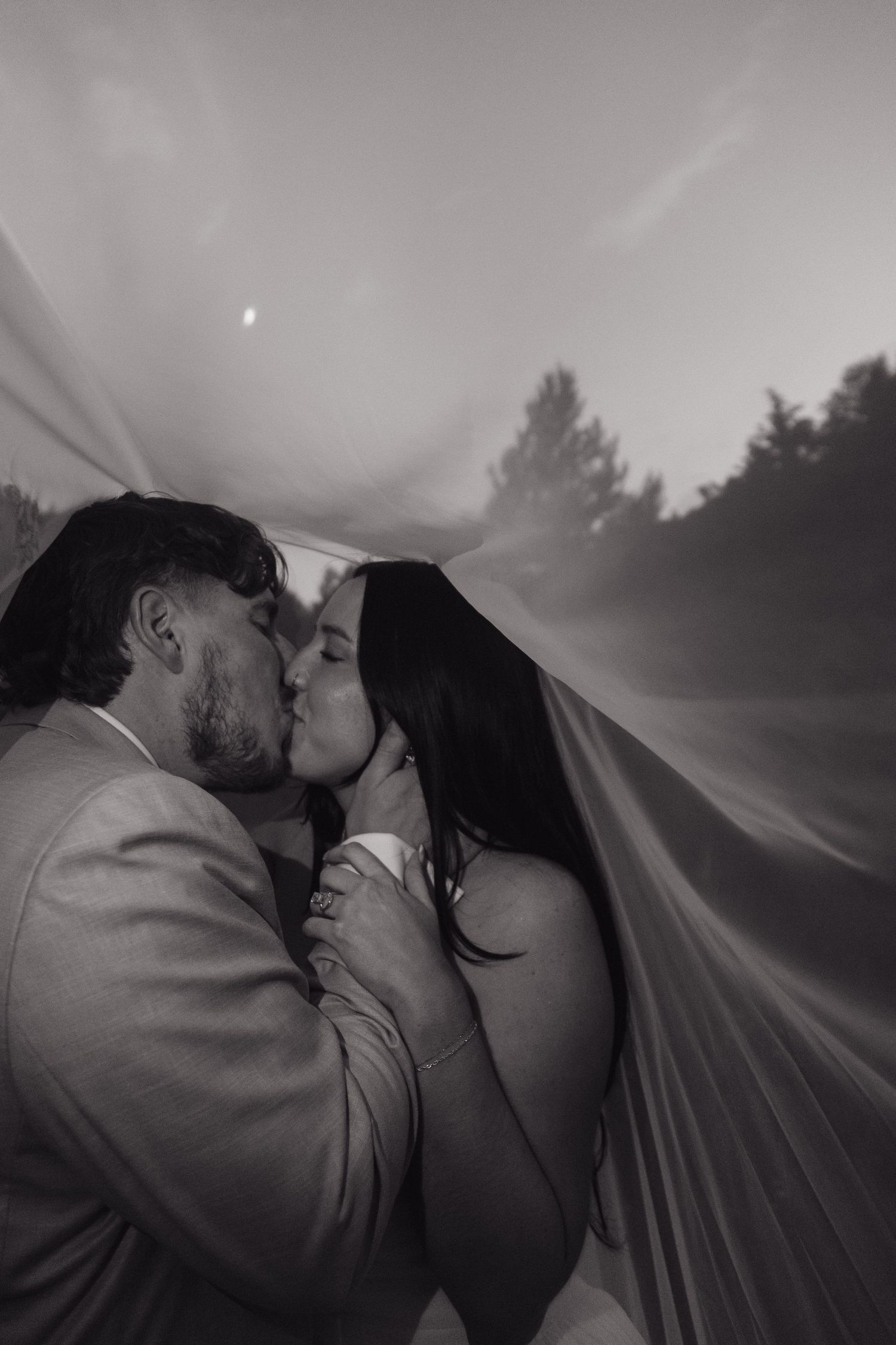 Black and white romantic photo of bride and groom kissing under flowing veil at dusk.