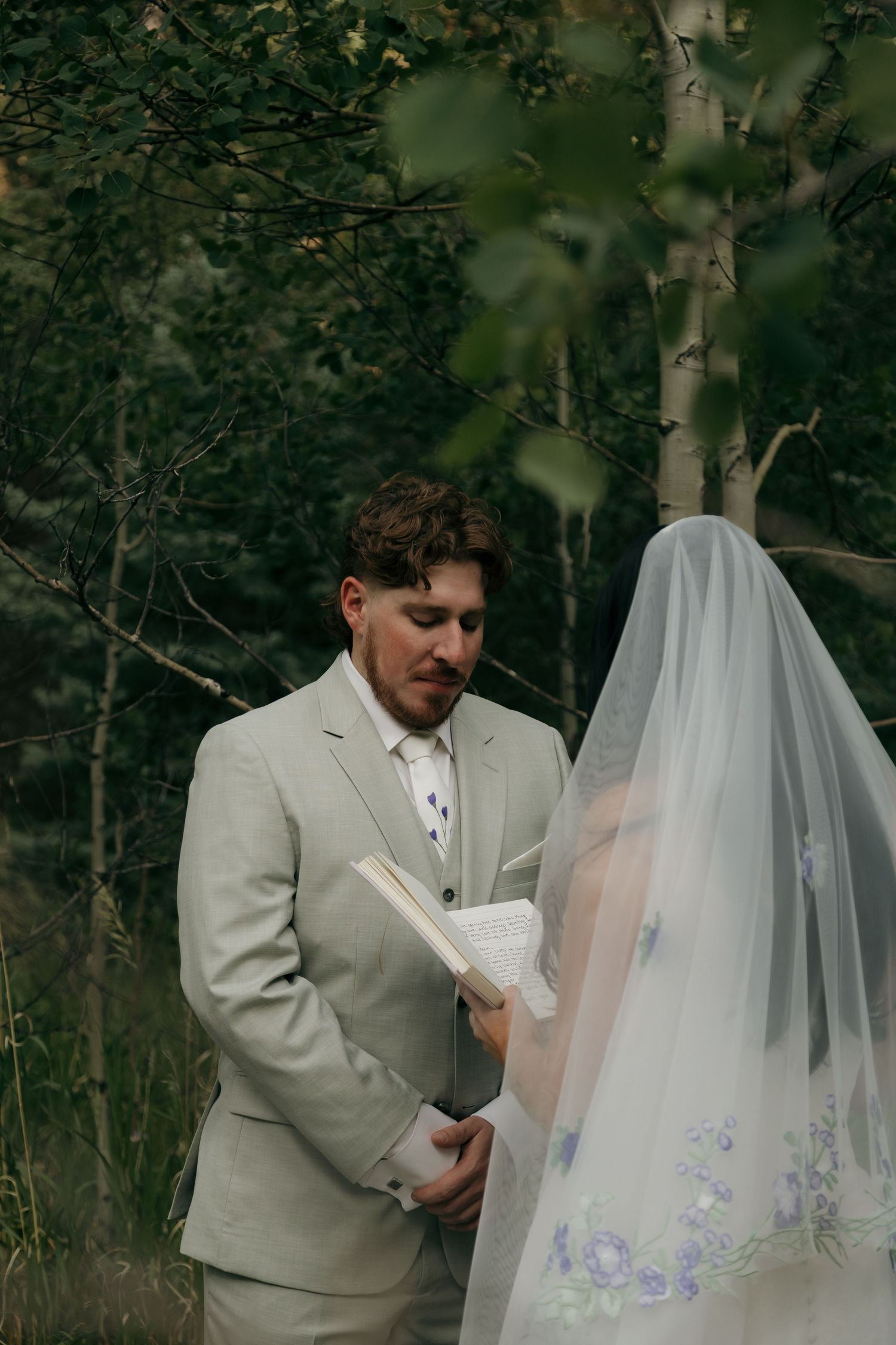 Groom in grey suit reads vows from a book to his veiled bride during an outdoor forest wedding.