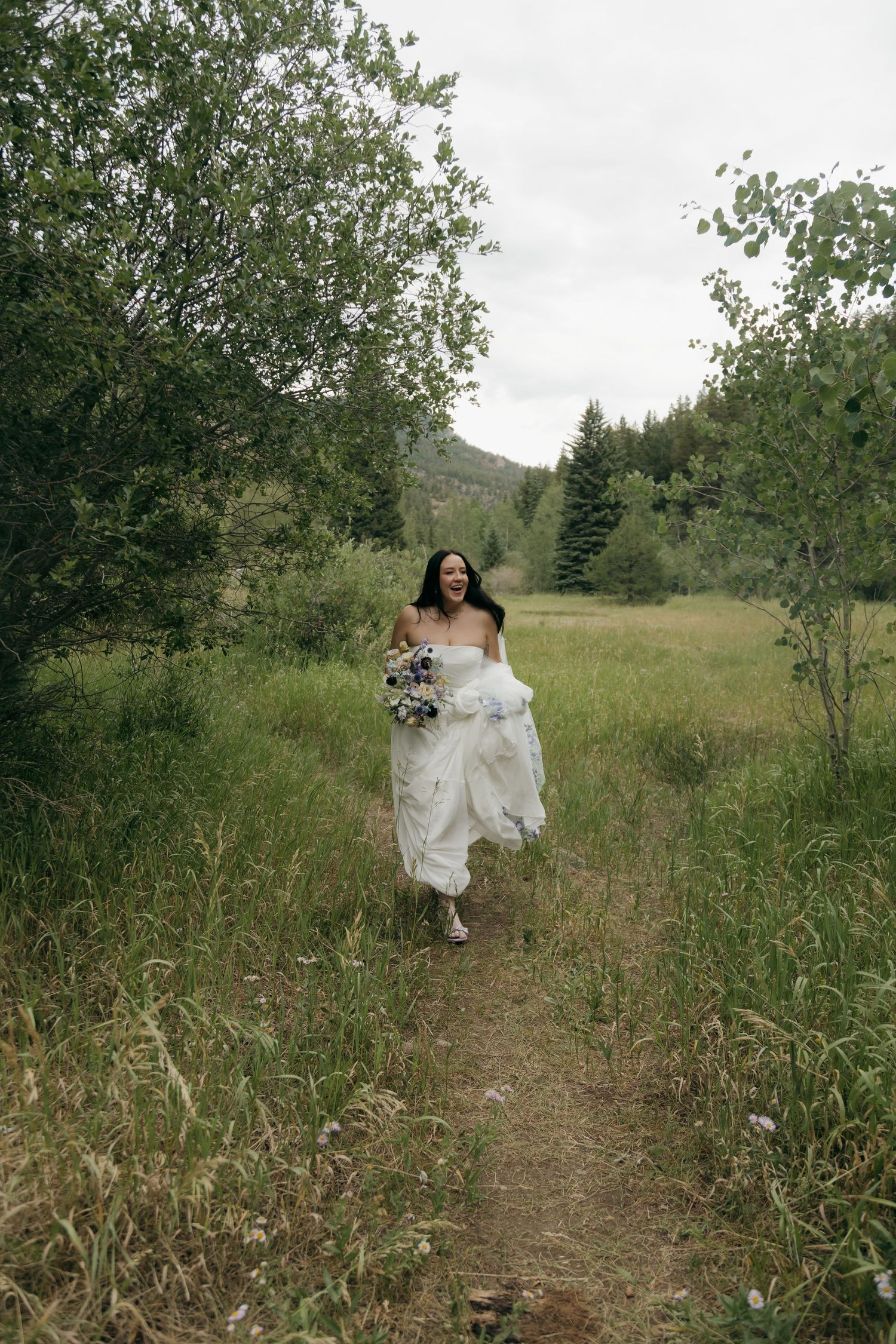 A joyful bride in a white gown runs along a forest path holding a bouquet, surrounded by lush green meadows.