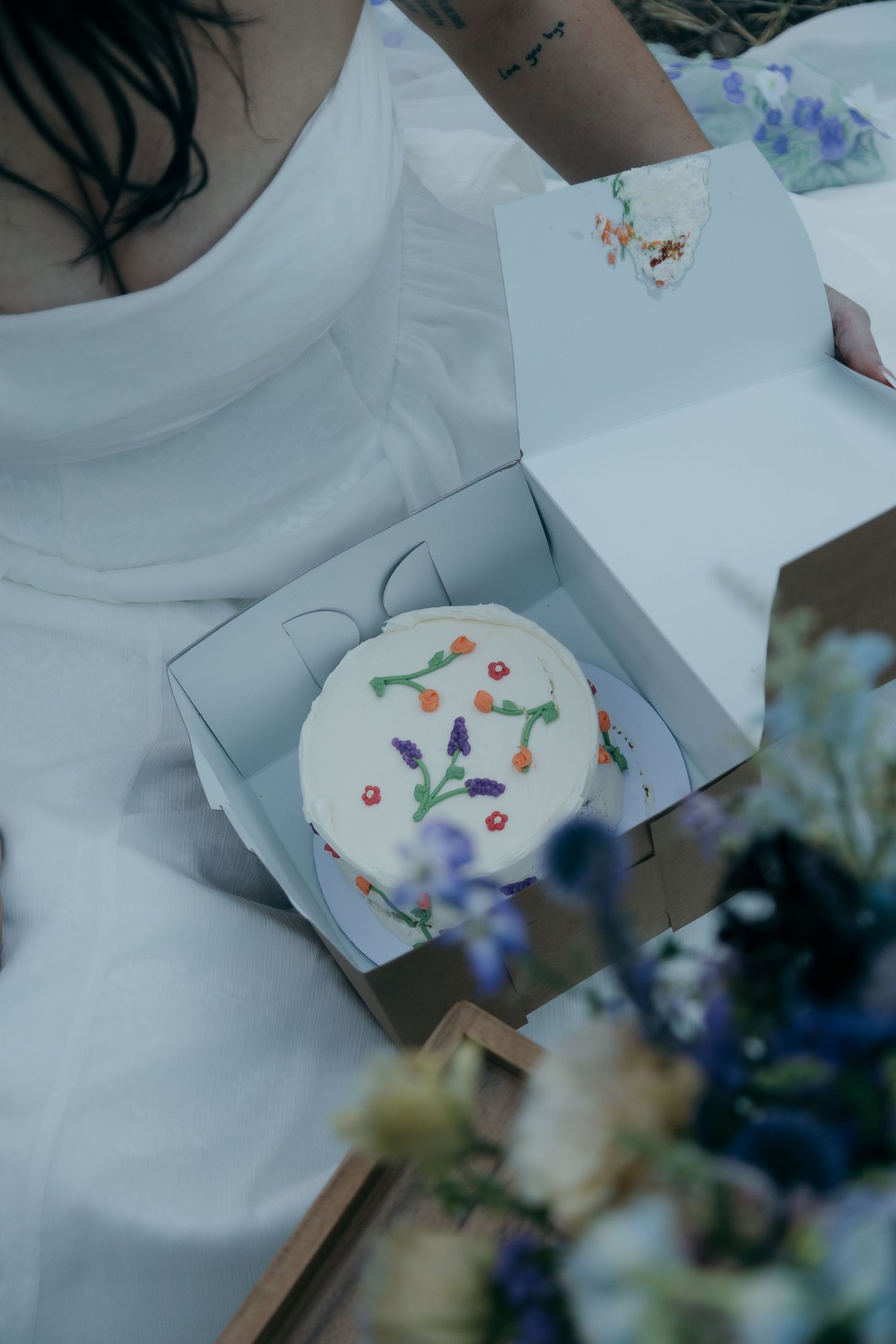 Bride holds open box with small white wedding cake decorated with colorful flowers.