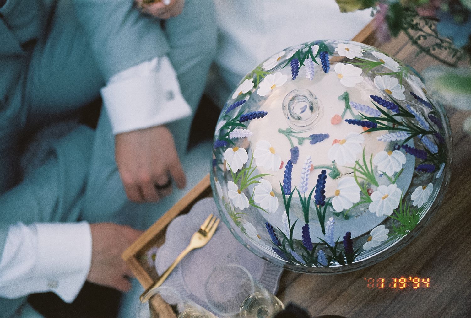 Overhead view of floral-painted wedding cake globe with white and blue flowers on picnic table.