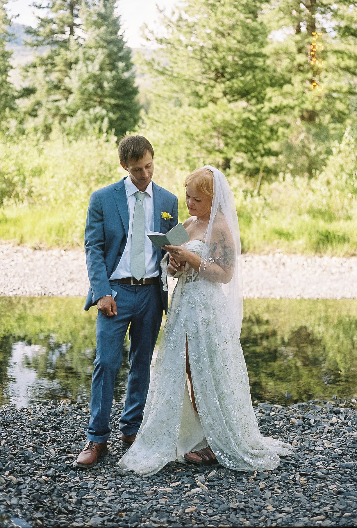 Bride in lace gown reads vows beside groom in blue suit on rocky riverbank surrounded by pine trees.