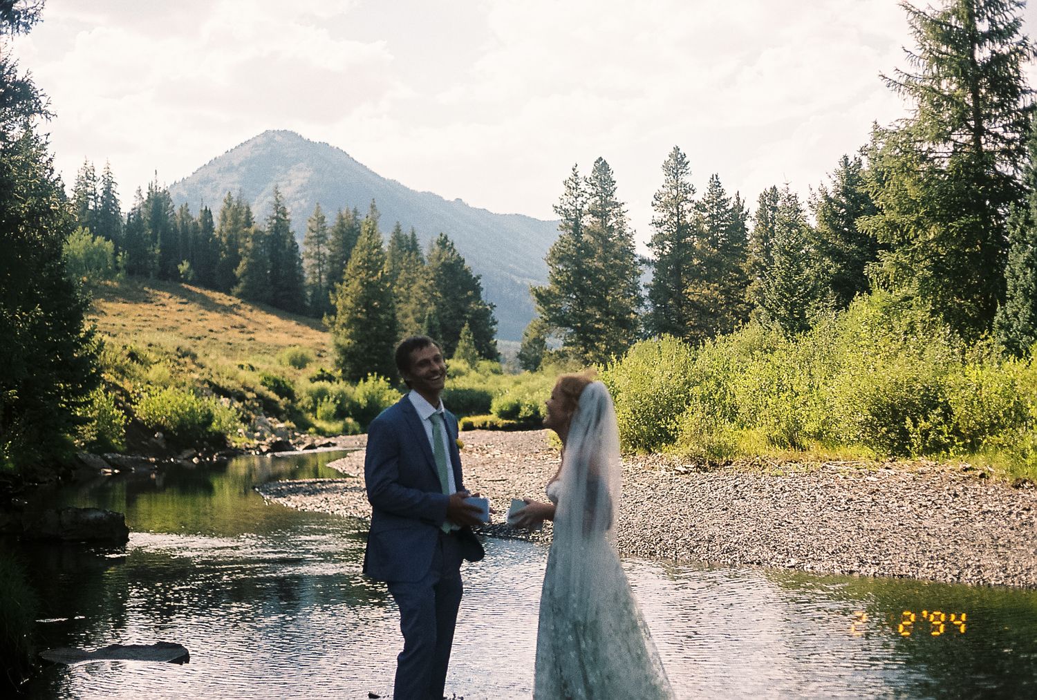 Groom in blue suit and veiled bride exchange vows beside a mountain stream surrounded by forest.