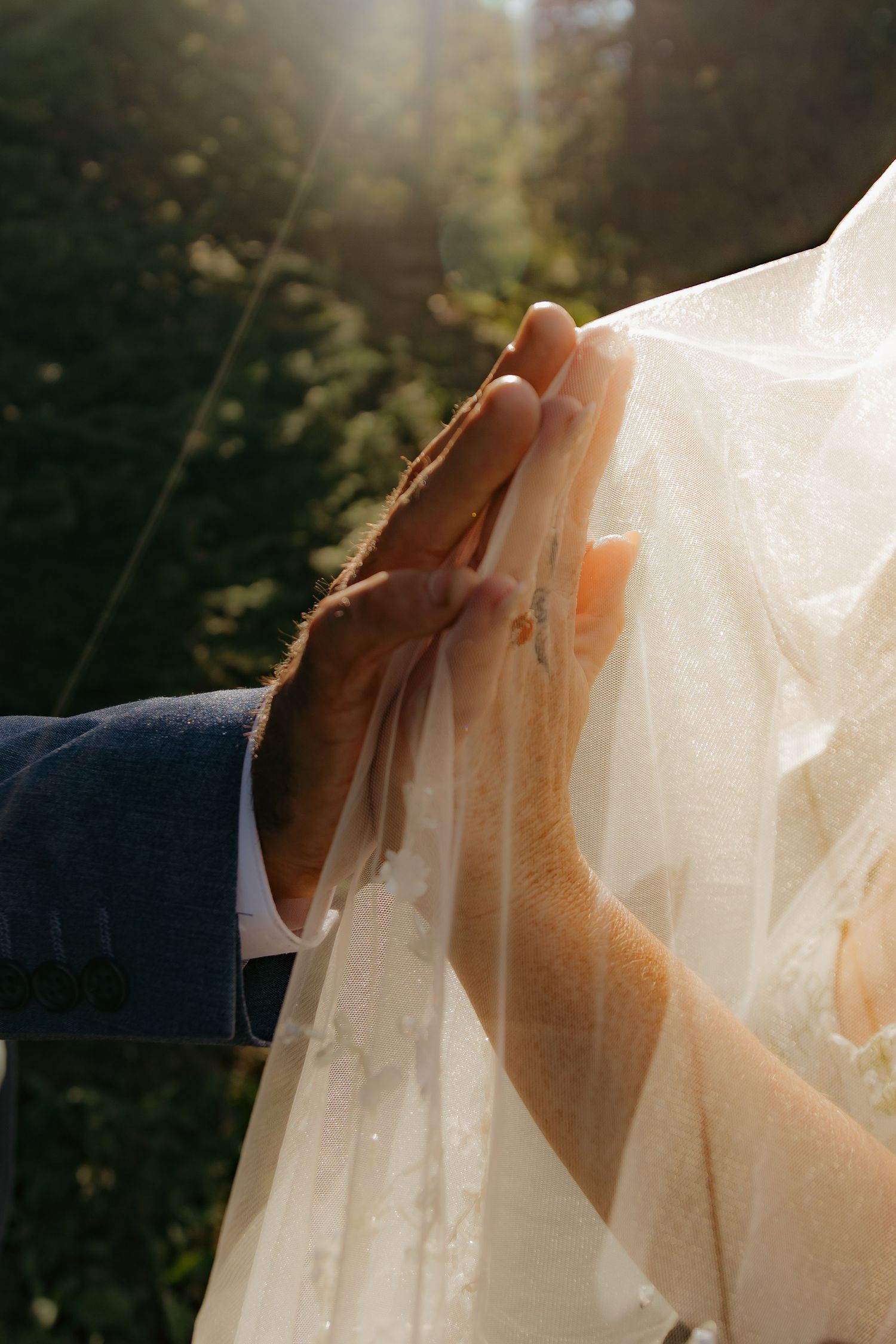 Groom's hand gently touching bride's lace veil in golden sunlight at outdoor wedding ceremony.